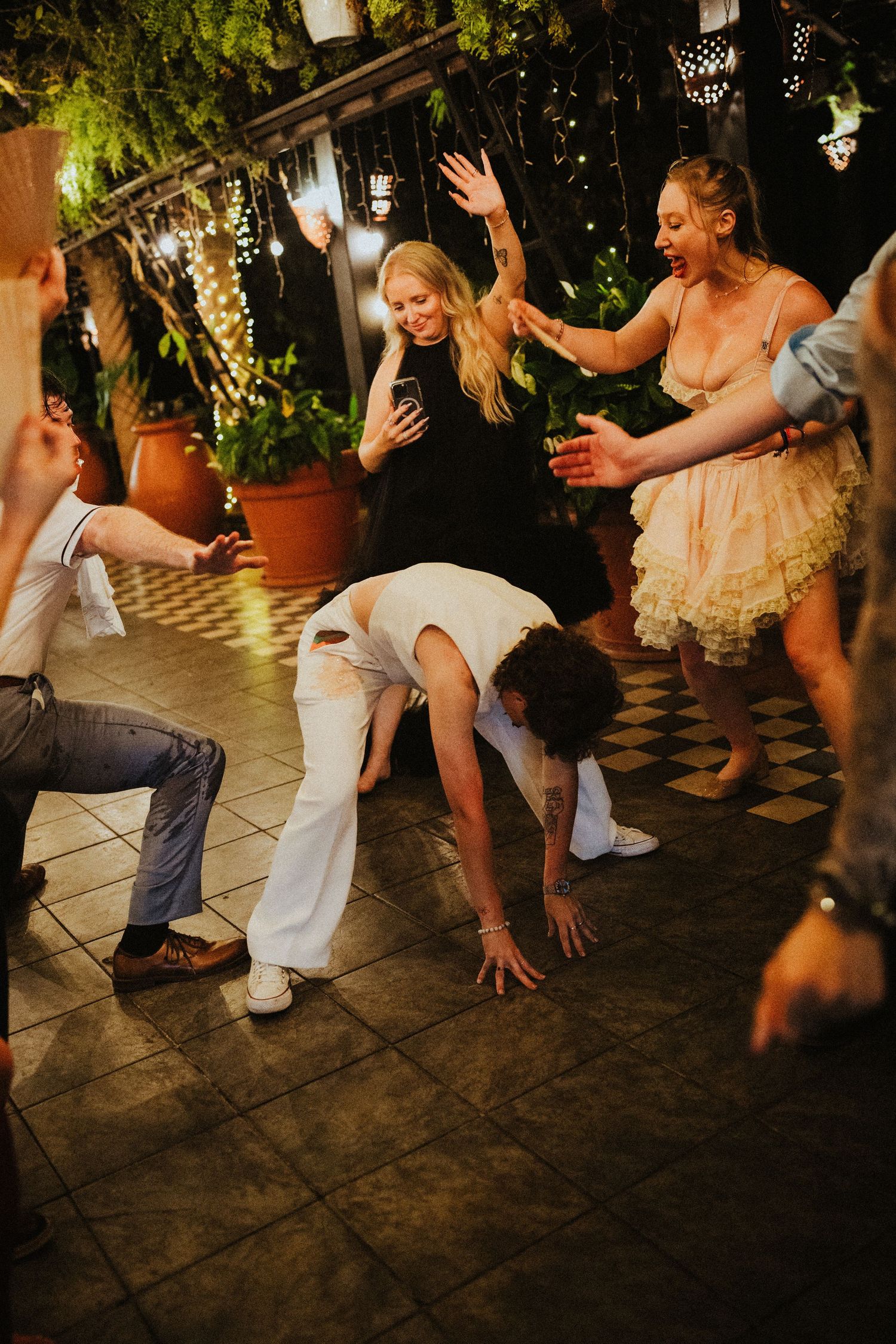 Guests dancing and celebrating at an outdoor evening party with string lights and tropical plants.