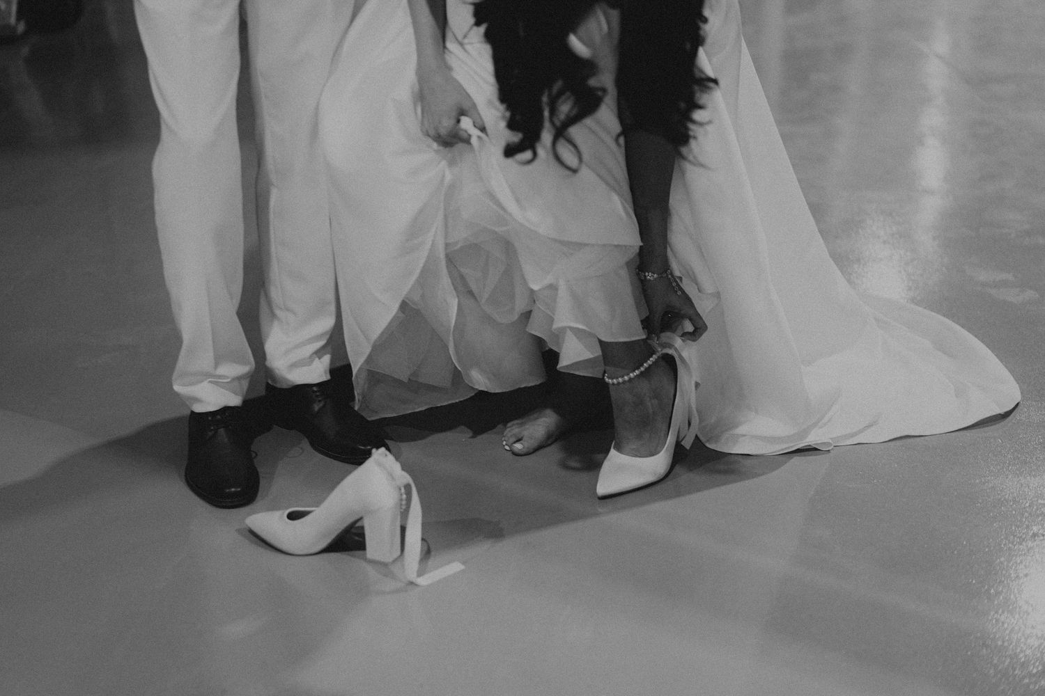 Black and white photo of bride and groom's feet, bride removing heels at wedding reception.
