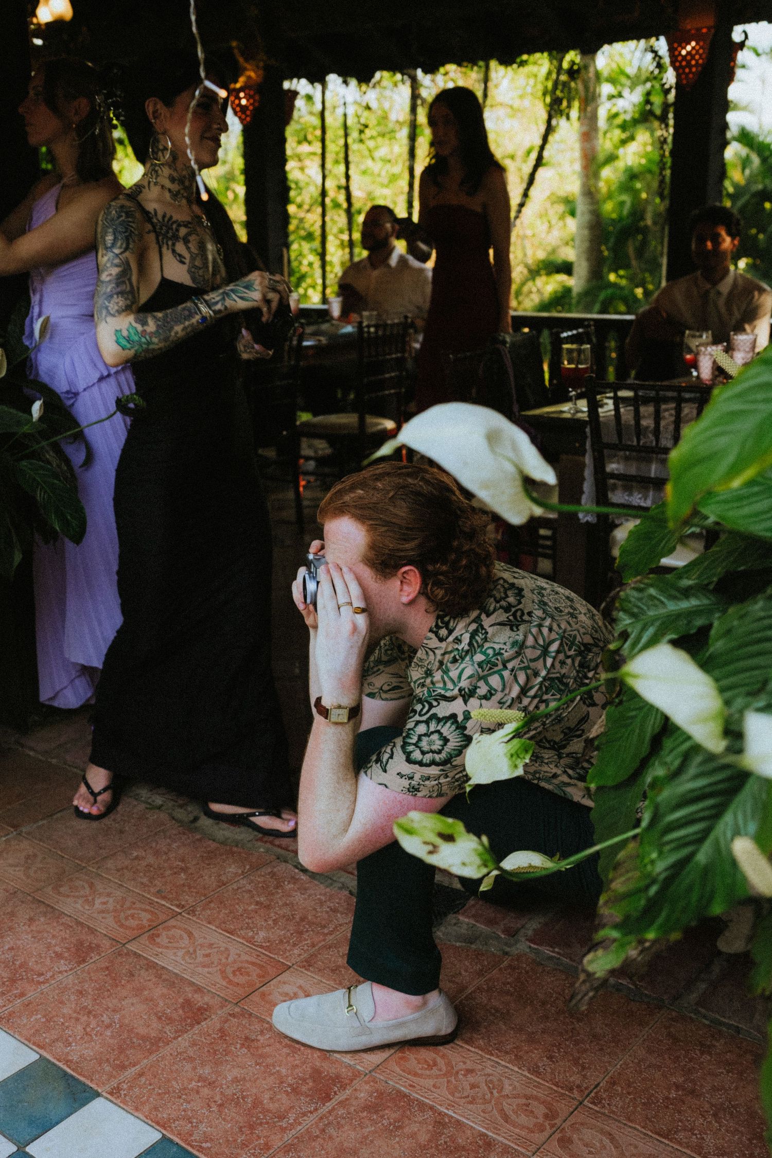 A photographer crouches among tropical plants to capture candid moments at an outdoor garden party event.