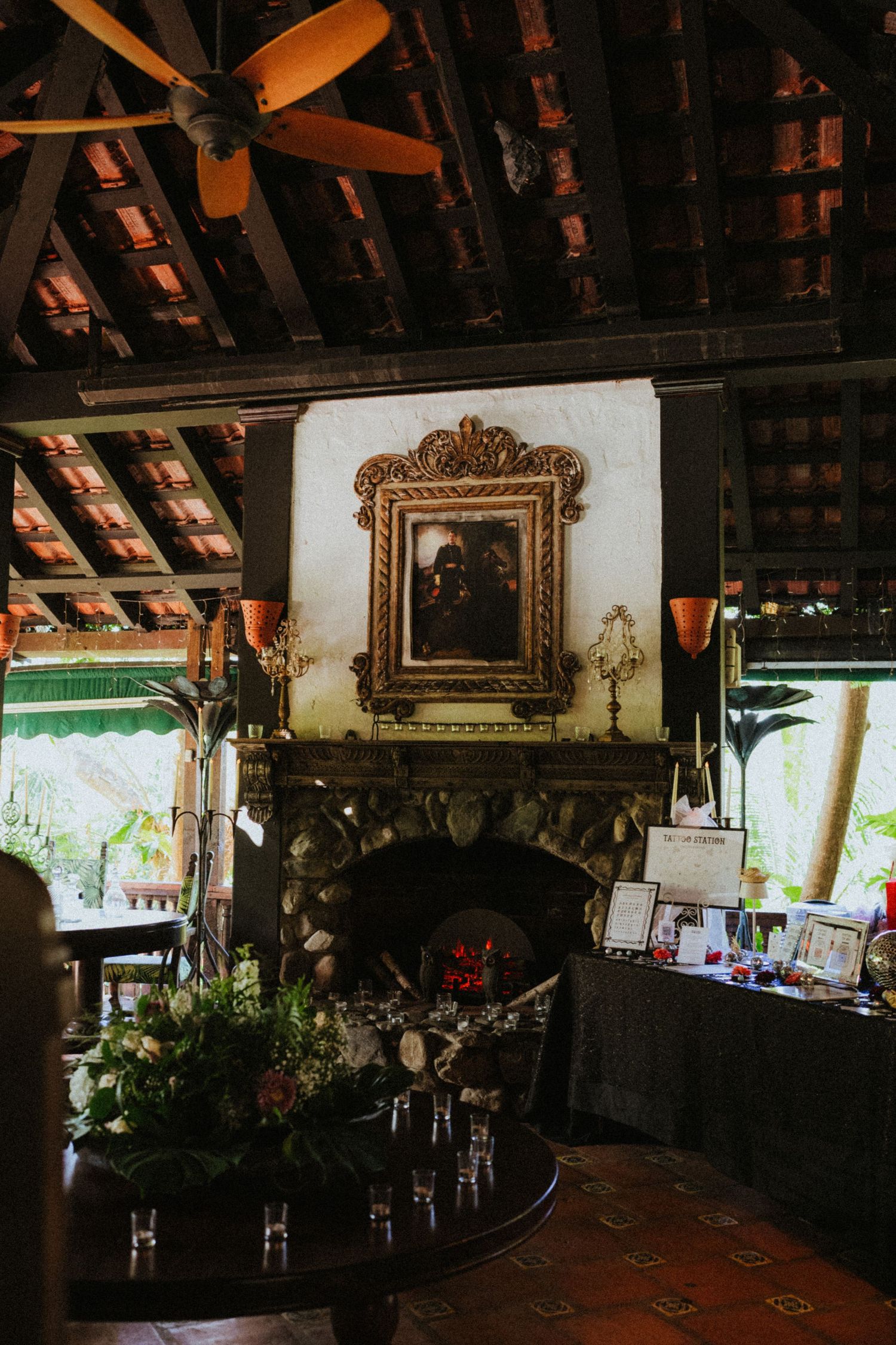 Rustic indoor venue with stone fireplace, ornate gold-framed painting, wooden ceiling, ceiling fan, and floral table decor.