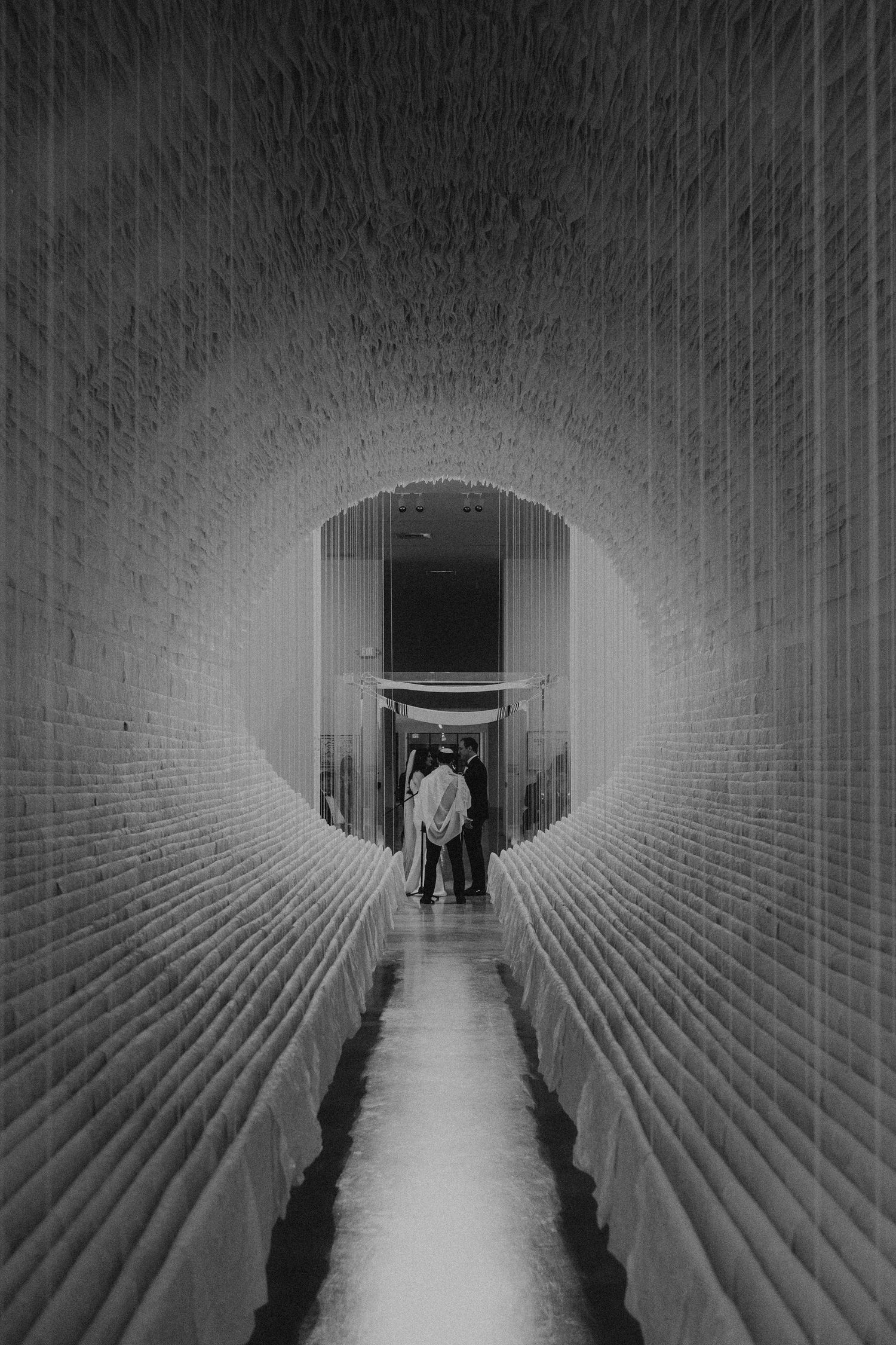 Black and white photo of a couple standing at the end of a narrow arched brick tunnel corridor with glowing light.