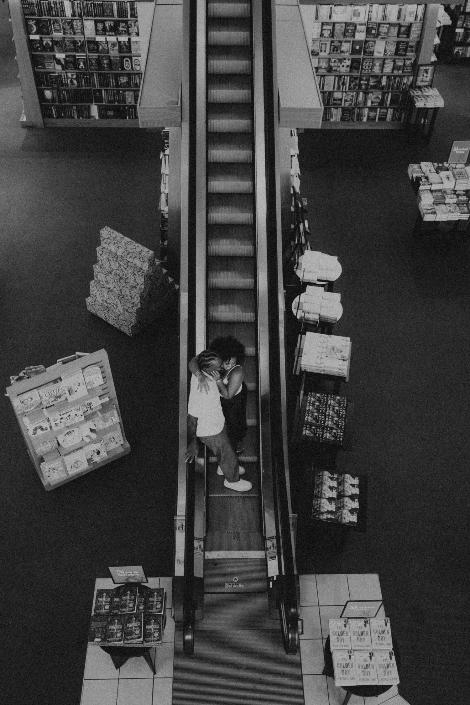 Aerial black and white view of two people on a bookstore escalator surrounded by shelves of books.