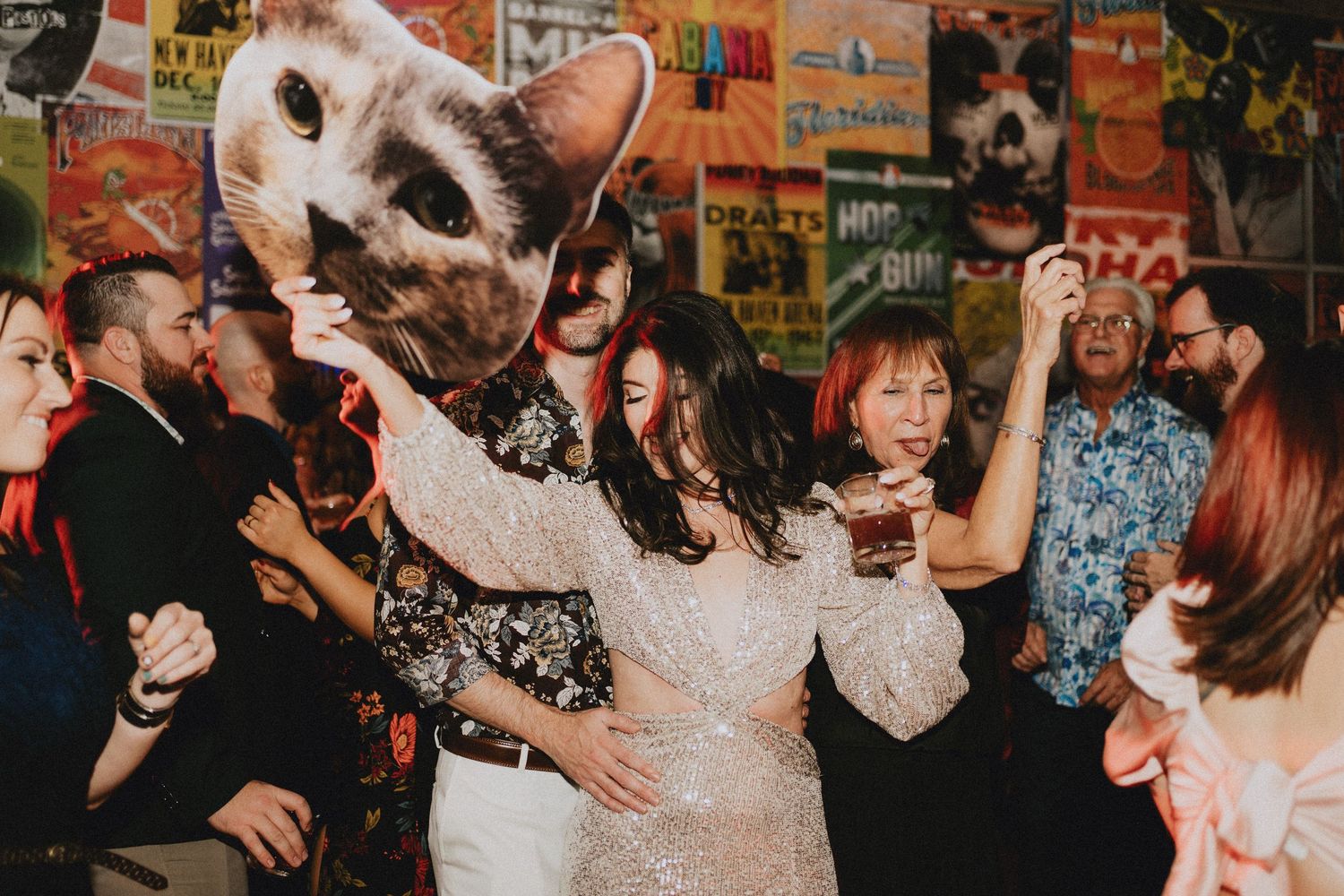 Wedding guests dancing and celebrating with a large cat face prop at a lively reception party.