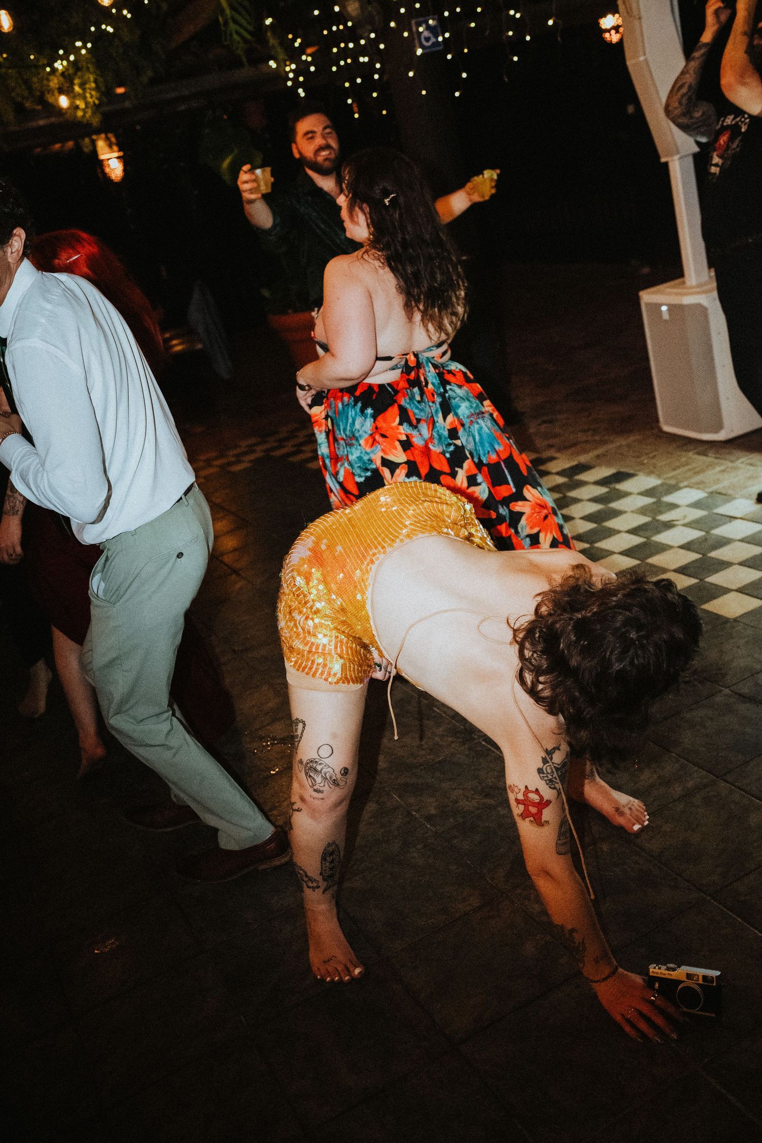 People dancing at a lively outdoor party, woman in gold sequin shorts and colorful dress bending down on checkered floor.