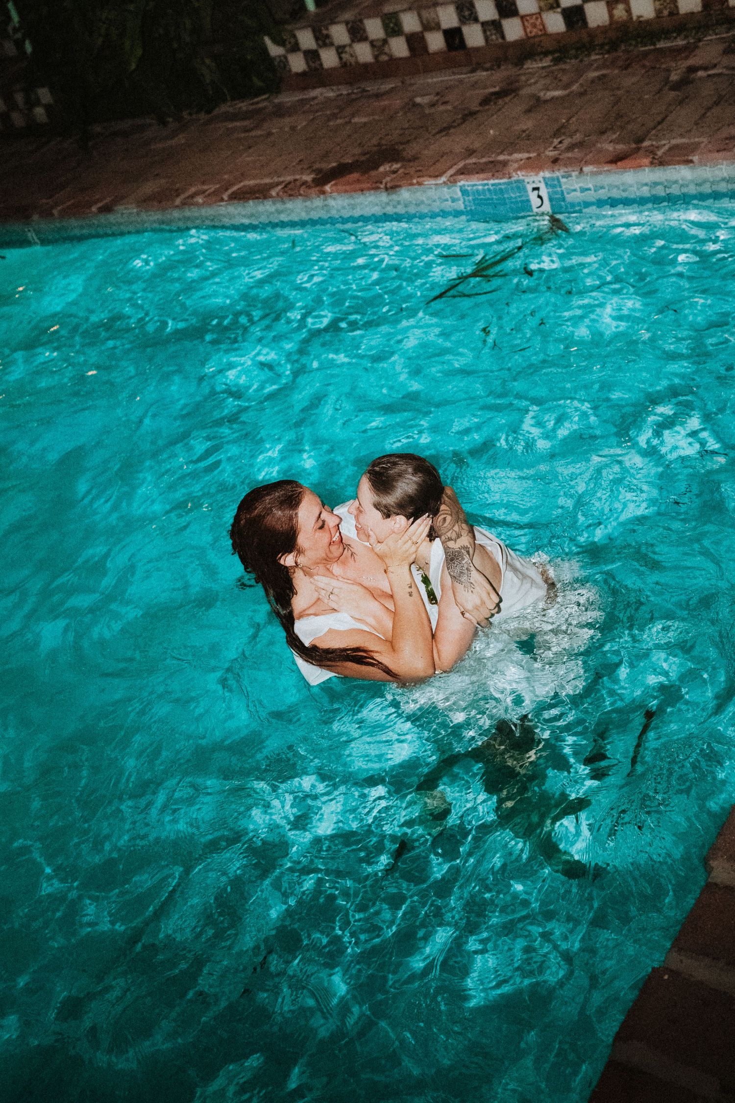 Two people sharing a romantic kiss while swimming in a turquoise pool at night.