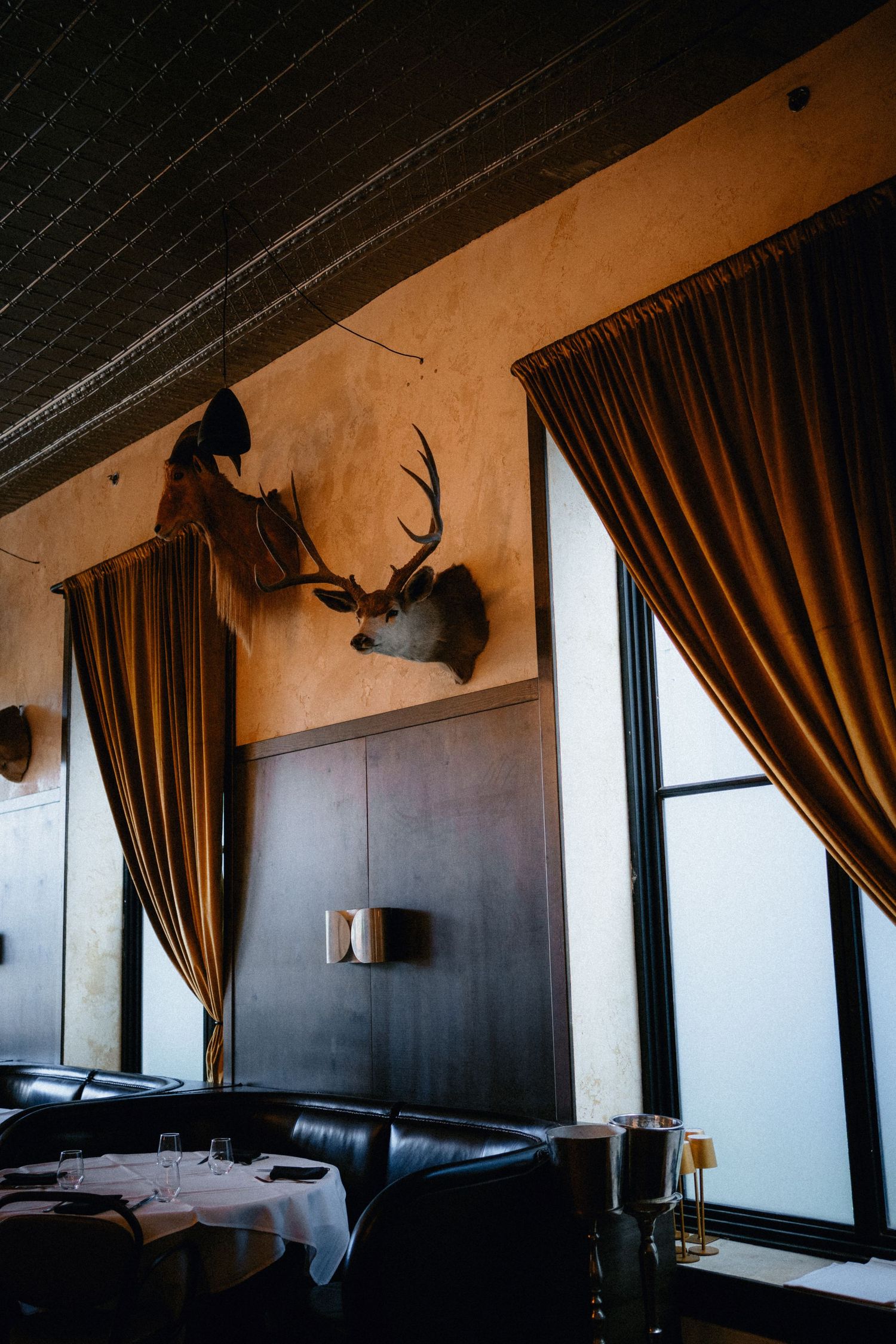 Rustic restaurant interior with mounted deer antlers, leather booths, white tablecloths, and warm amber curtains framing large windows.