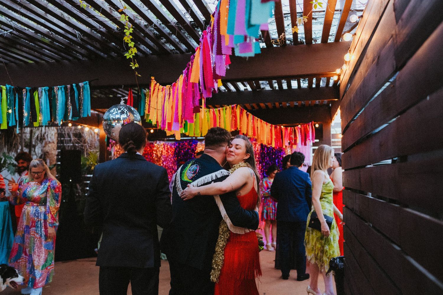 Couple embracing under colorful hanging fringe decorations at a festive outdoor wedding celebration.