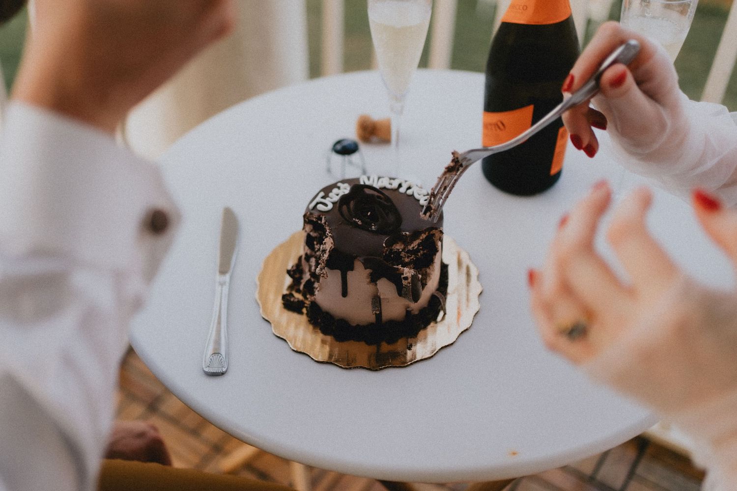 Two people sharing a chocolate drip cake on a white table with a bottle of Veuve Clicquot champagne.