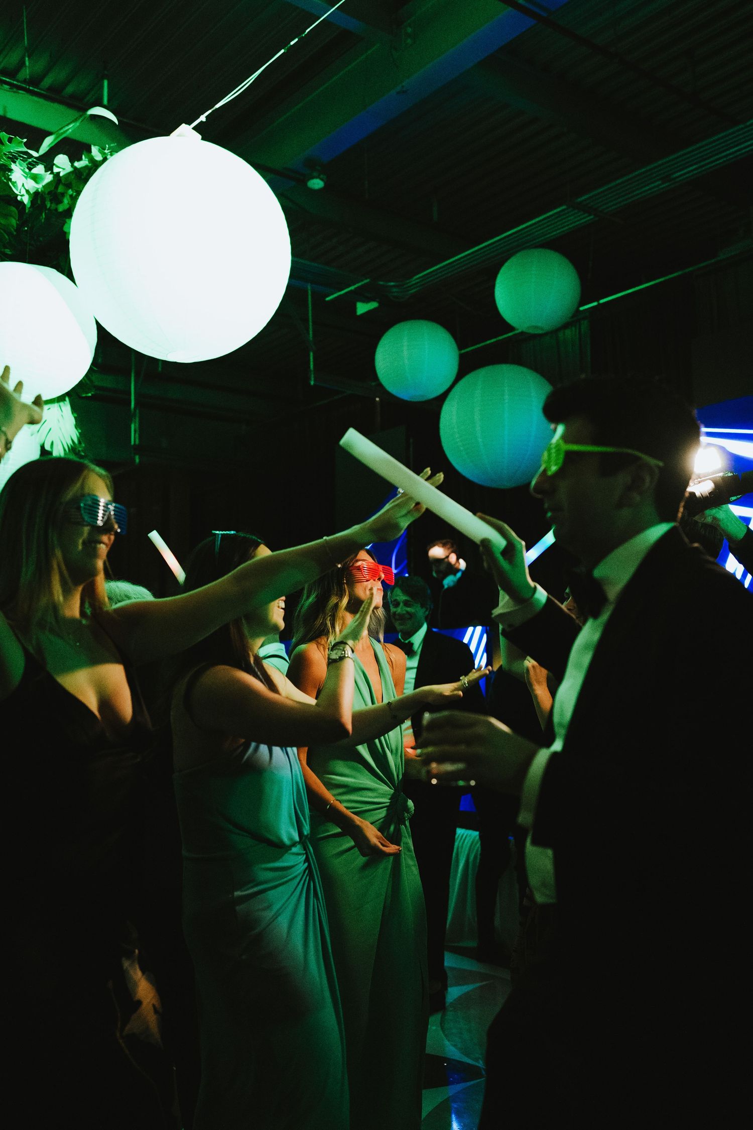 Guests dancing at a party under glowing white balloon lanterns in a dimly lit venue with green lighting.