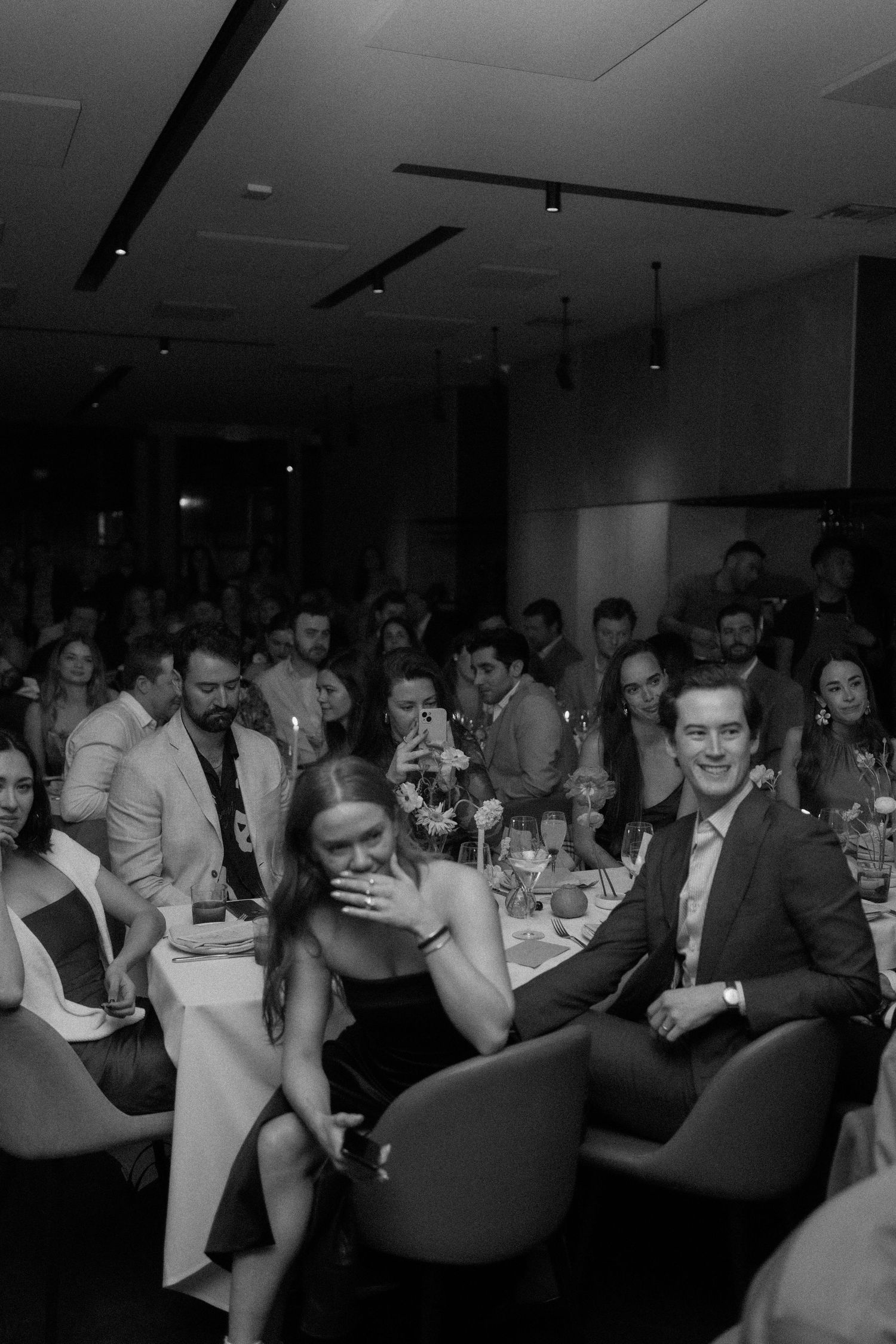 Wedding reception guests laughing and smiling at dinner tables in a crowded venue, black and white.