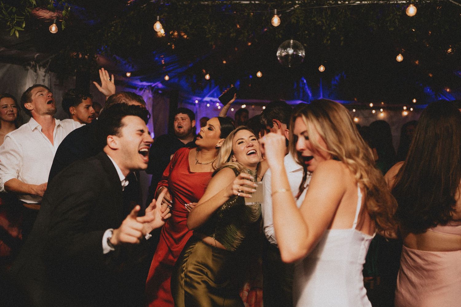 Guests dancing and singing joyfully at lively wedding reception with string lights and disco ball.