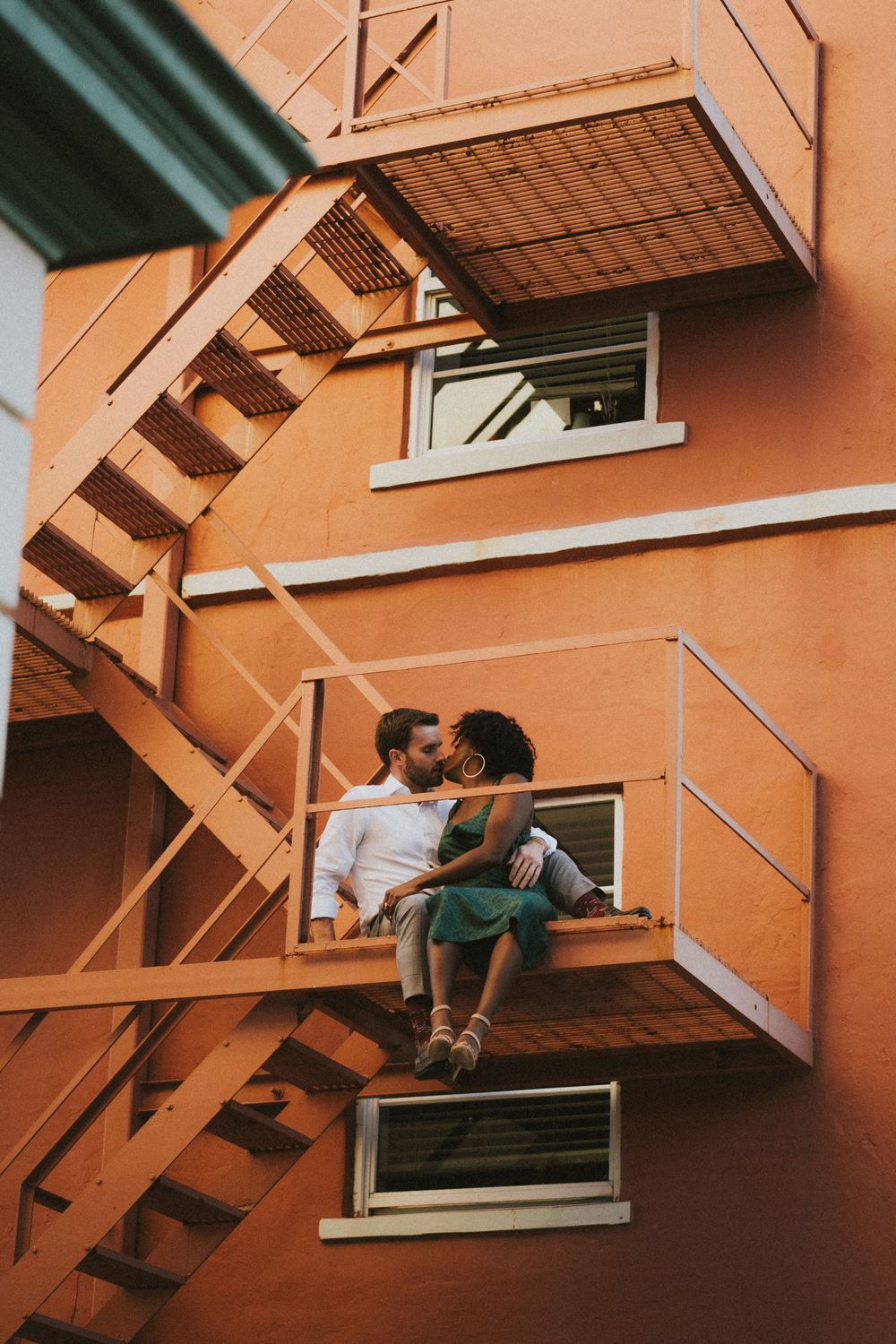 Romantic moment captured in series on outdoor metal fire escape with peachy colored wall background.