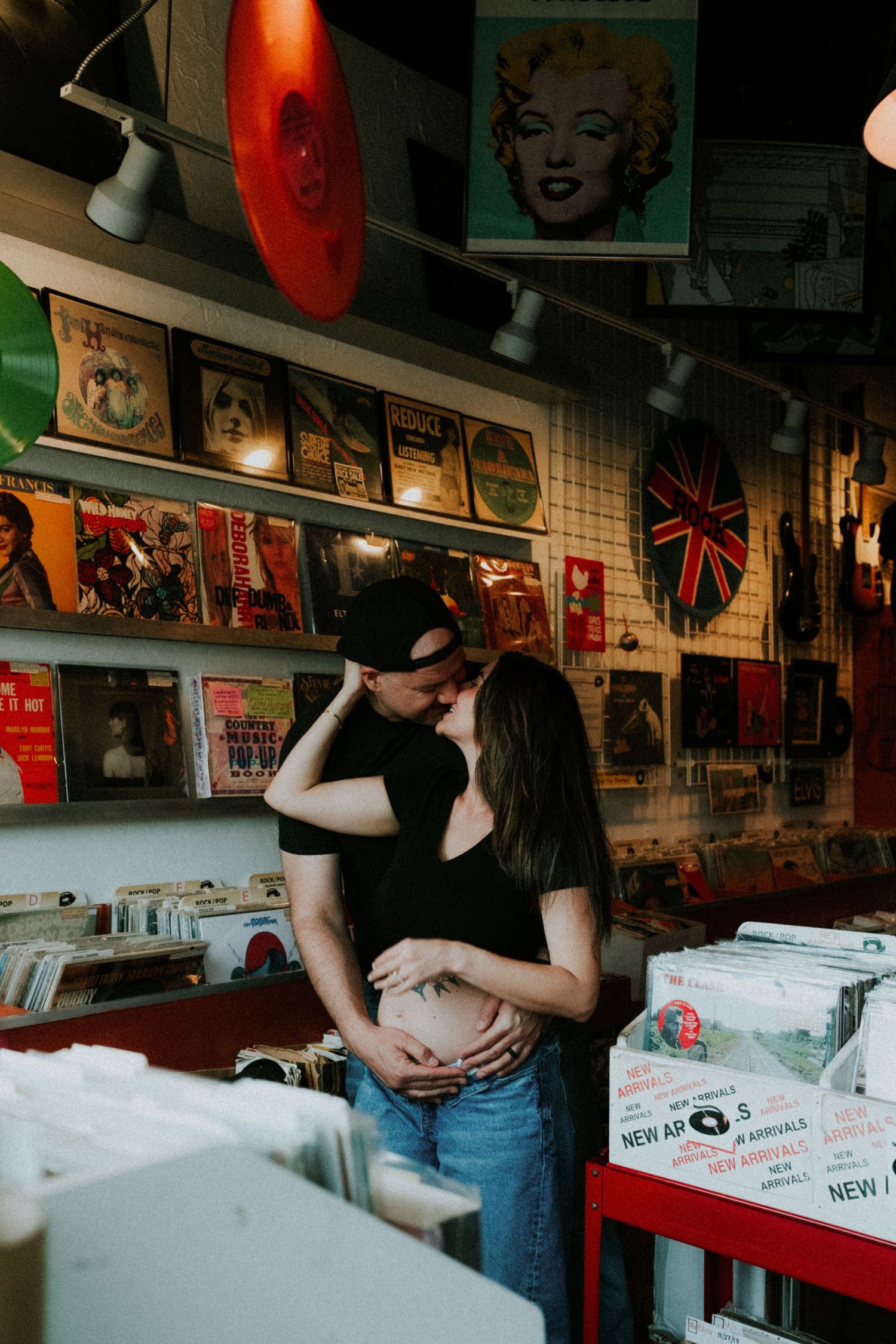 Couple sharing a tender moment in a vintage vinyl record shop, with the woman cradling her baby bump.