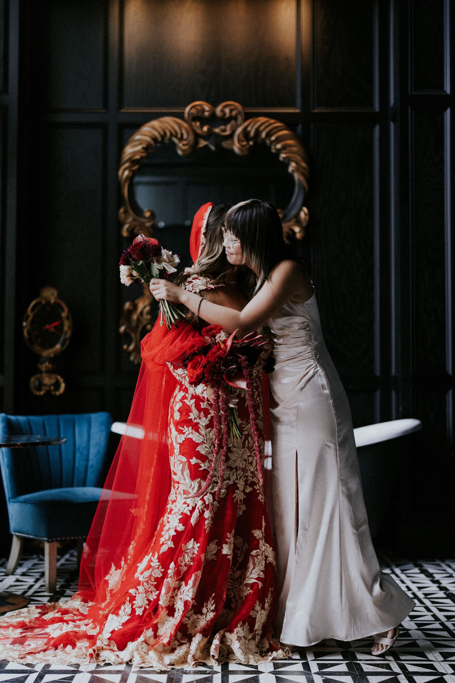 Bride in a stunning red and gold lace wedding dress embracing bridesmaid in a dark elegant venue.