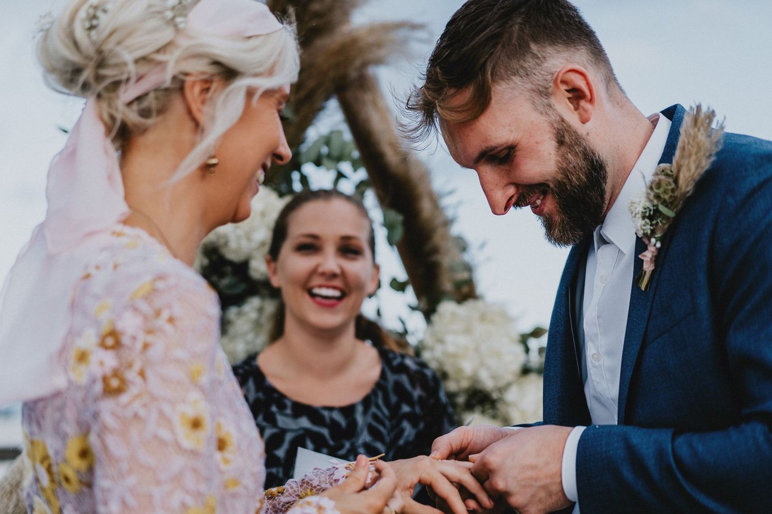 Bride and groom exchanging rings during outdoor wedding ceremony while smiling officiant watches joyfully.
