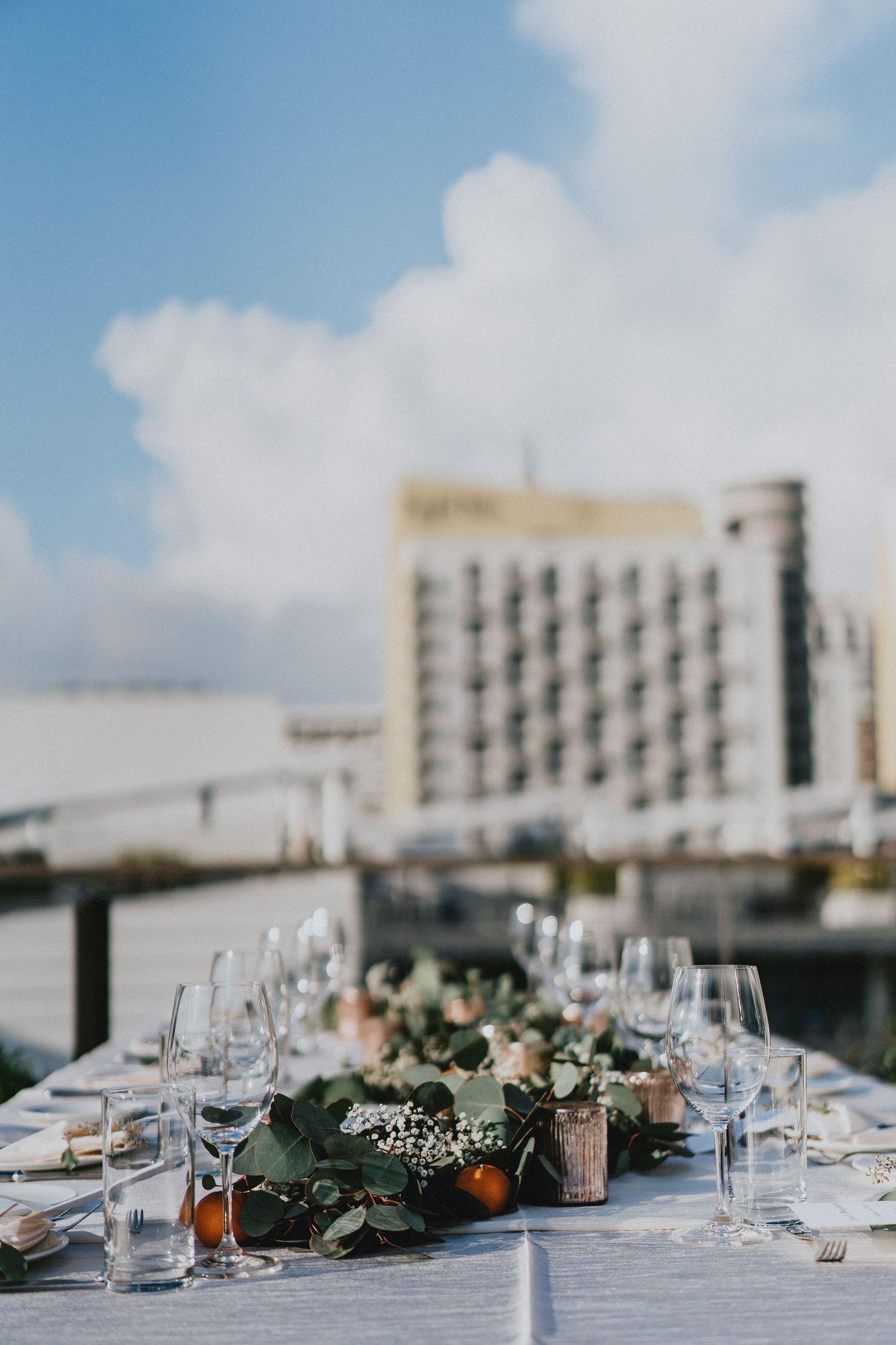 Elegant outdoor rooftop wedding reception table with floral greenery centerpiece, glassware, and city building backdrop.