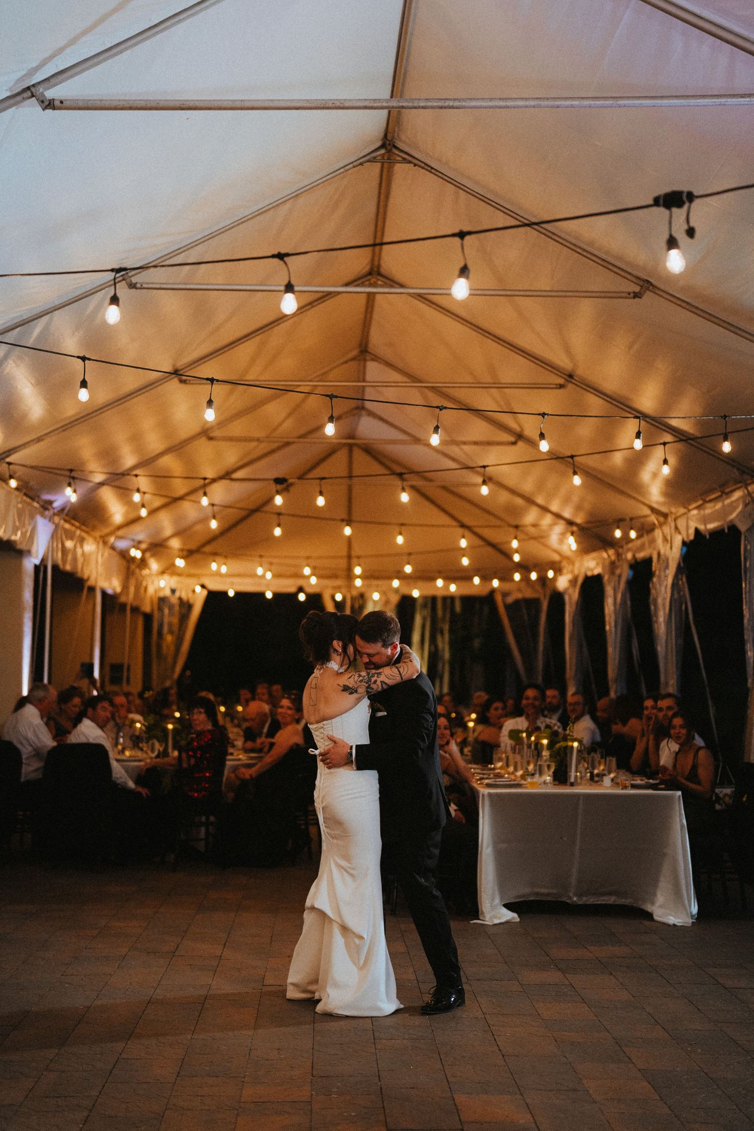 Bride and groom share their first dance under a white tent adorned with warm string lights at their wedding reception.