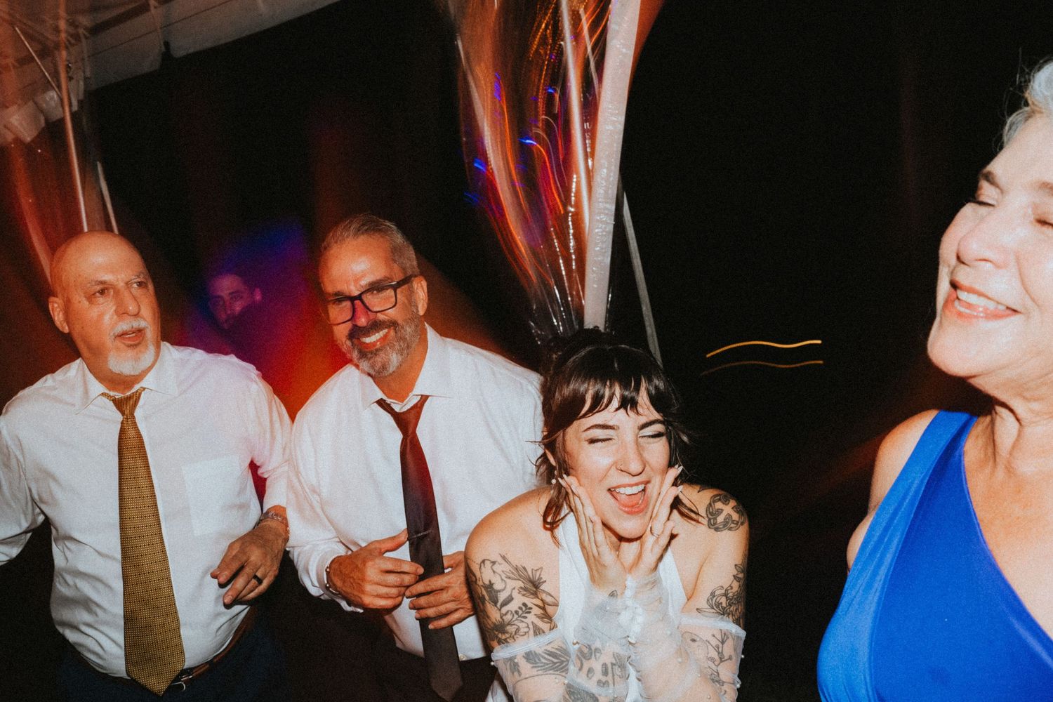Joyful wedding reception guests dancing and laughing together, bride in white dress with tattoos celebrating with family.