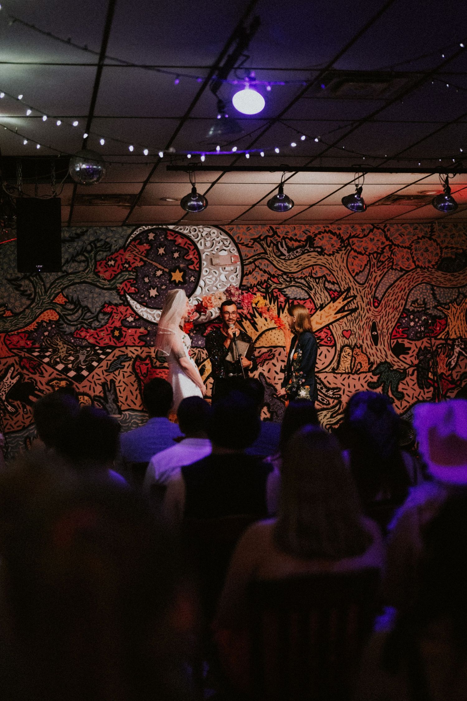 Crowd gathered at a vibrant venue with colorful mural backdrop and string lights during a lively indoor event.