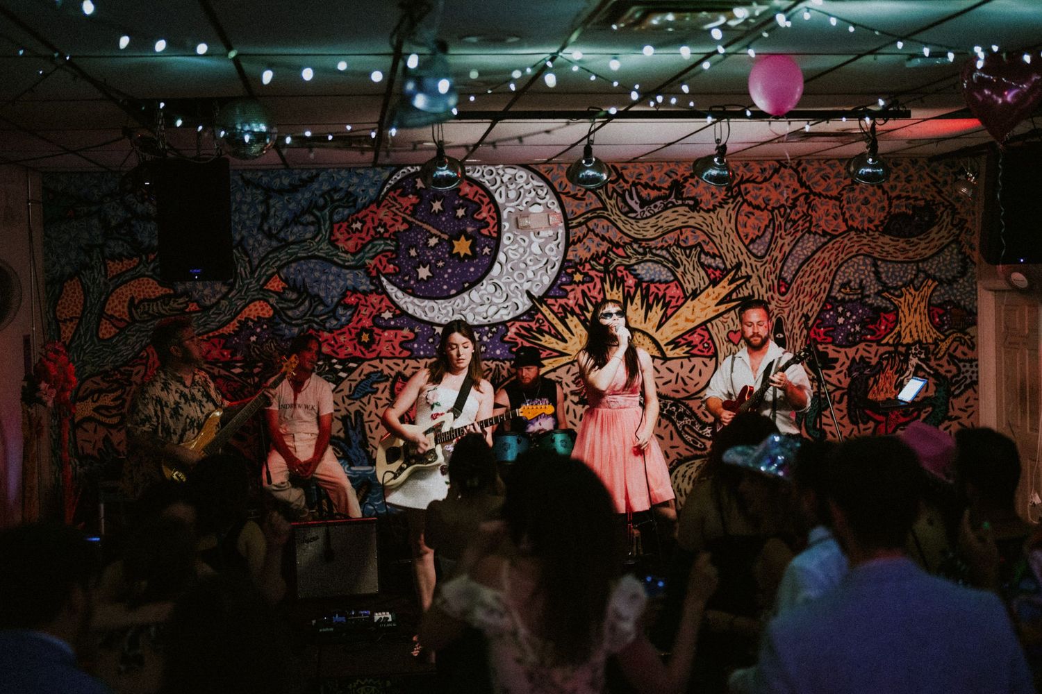 Live band performing on stage with colorful psychedelic mural backdrop featuring moon and sun artwork.