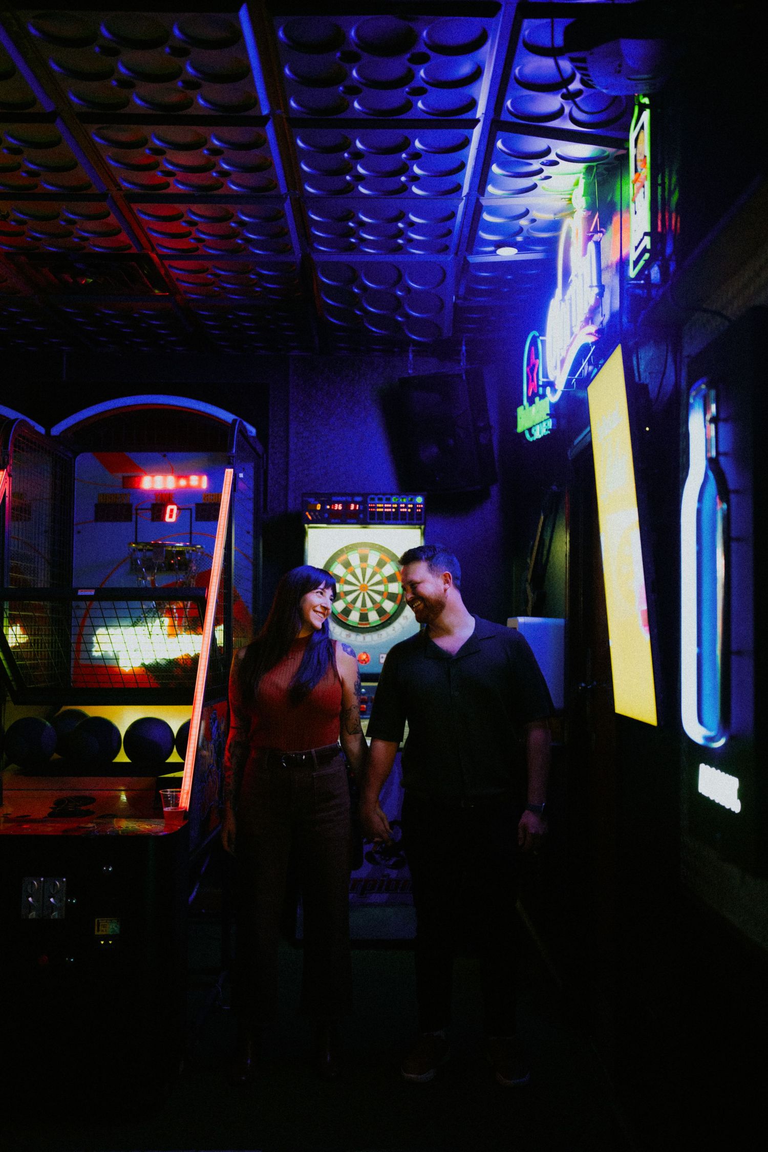 Two people stand in a dimly lit arcade with neon lights and a dartboard illuminated in the background.
