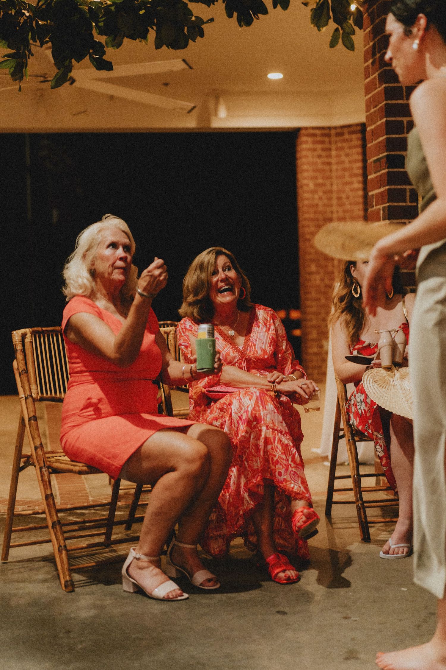 Wedding party members in coral dresses sit on wooden chairs during a candid reception moment.