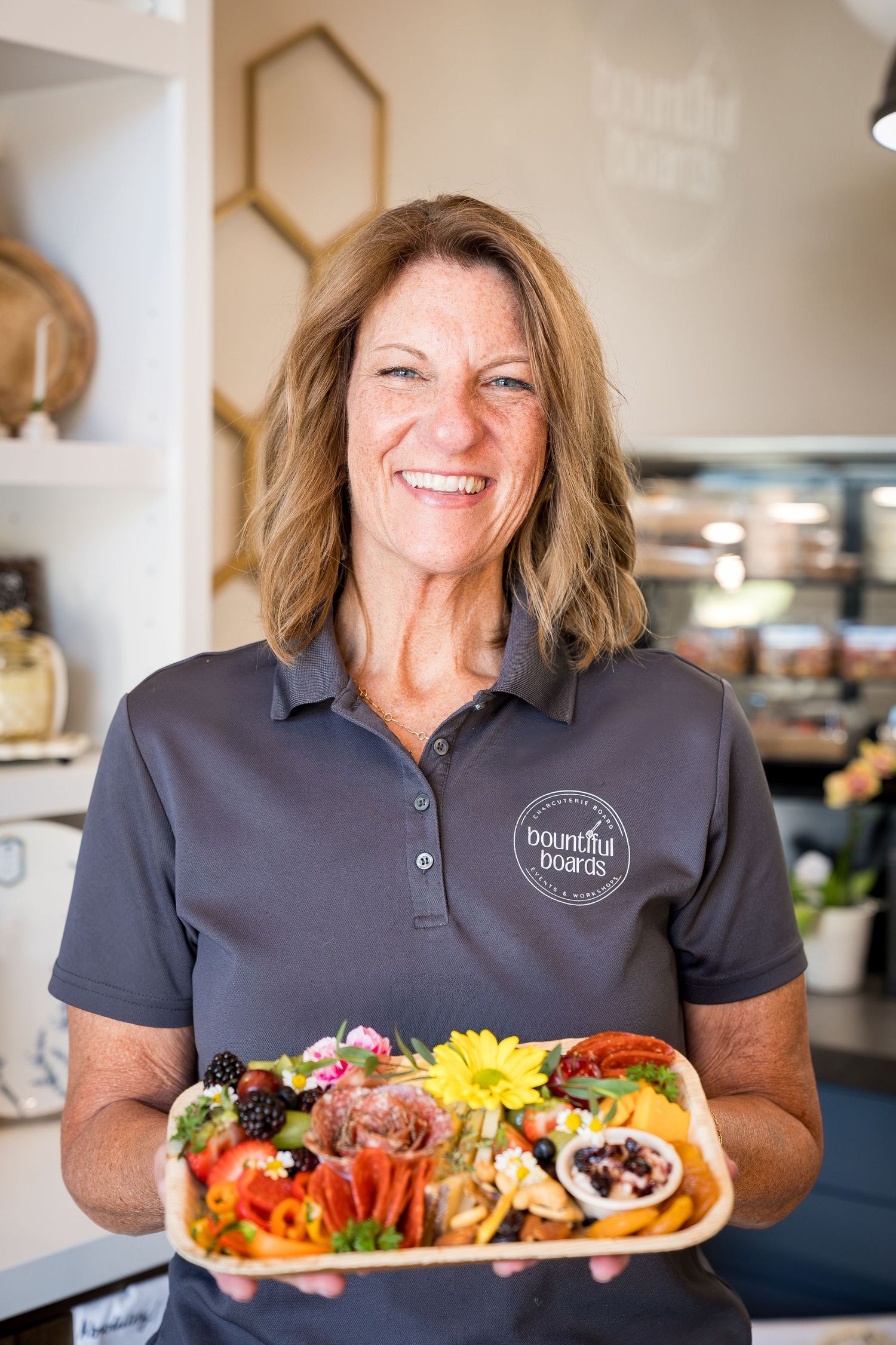 Smiling woman holding a colorful charcuterie board wearing a Bountiful Boards logo shirt in a cafe.