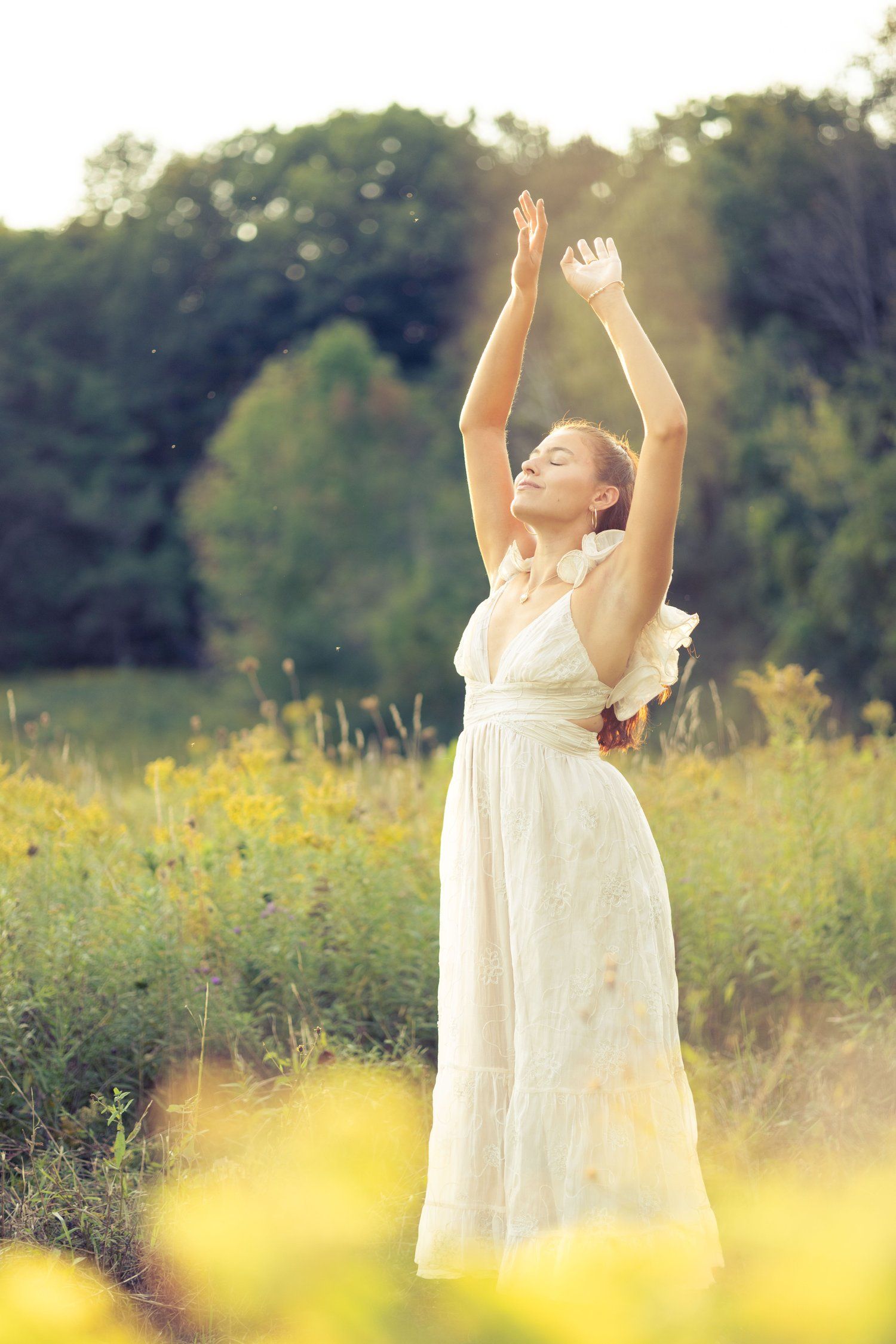 Woman in a white dress standing in a sunlit field of wildflowers with arms raised and eyes closed, surrounded by greenery.