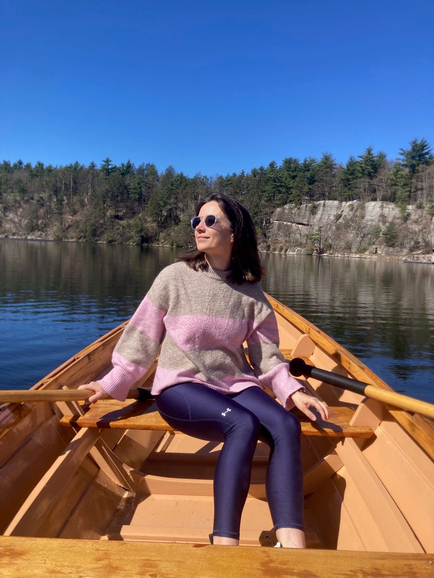Woman wearing sunglasses and a pink and beige sweater sitting in a wooden rowboat on a calm lake, surrounded by trees and rocky cliffs