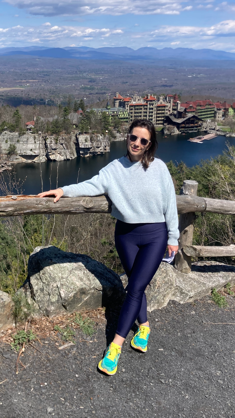 Woman wearing sunglasses, a light blue sweater, navy leggings, and bright yellow sneakers standing and leaning on a wooden fence with a scen