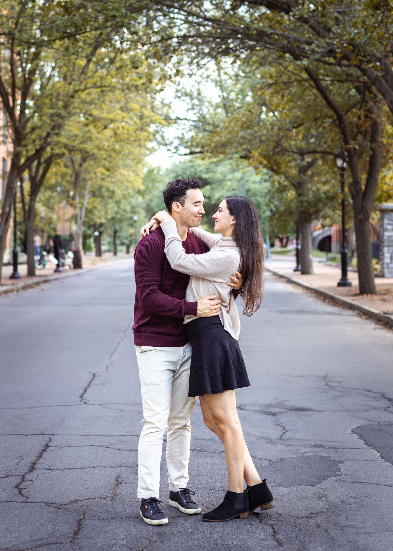 Couple embracing and smiling in the middle of a tree-lined street with autumn leaves and a calm, romantic atmosphere.