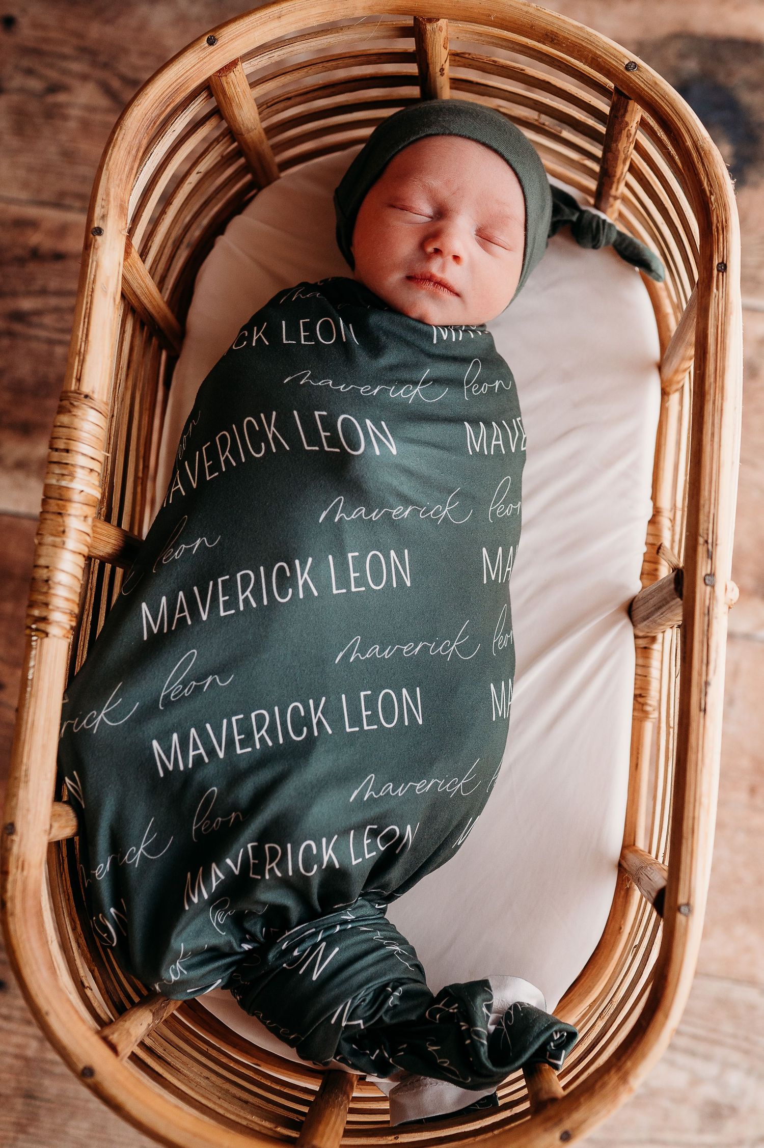 Newborn baby swaddled in custom printed blanket resting in woven basket during professional photo session.