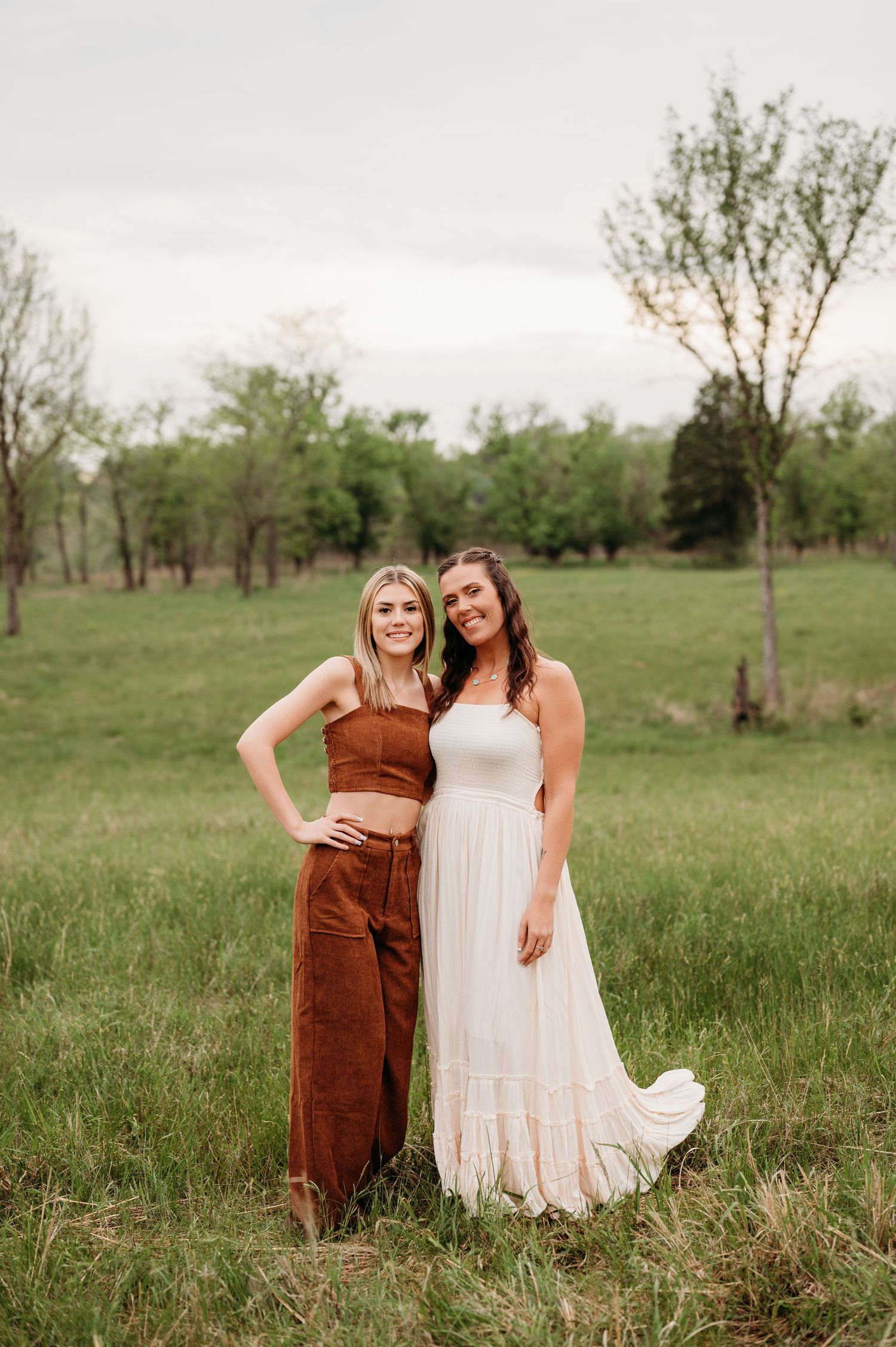 Two people in white and brown dresses pose together in a grassy field during a spring photoshoot.