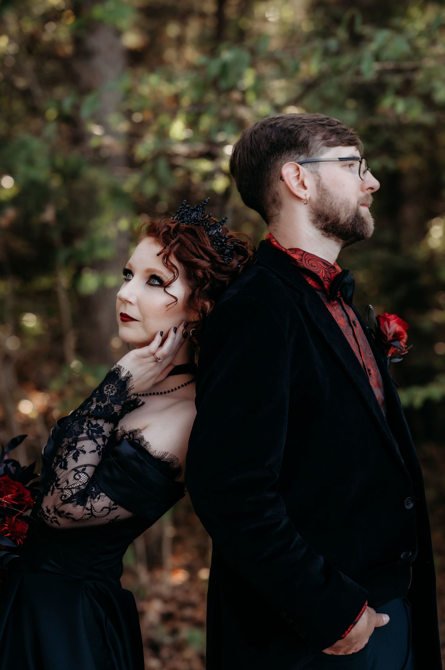 A couple in gothic wedding attire poses dramatically in a forest setting with black and red color scheme.