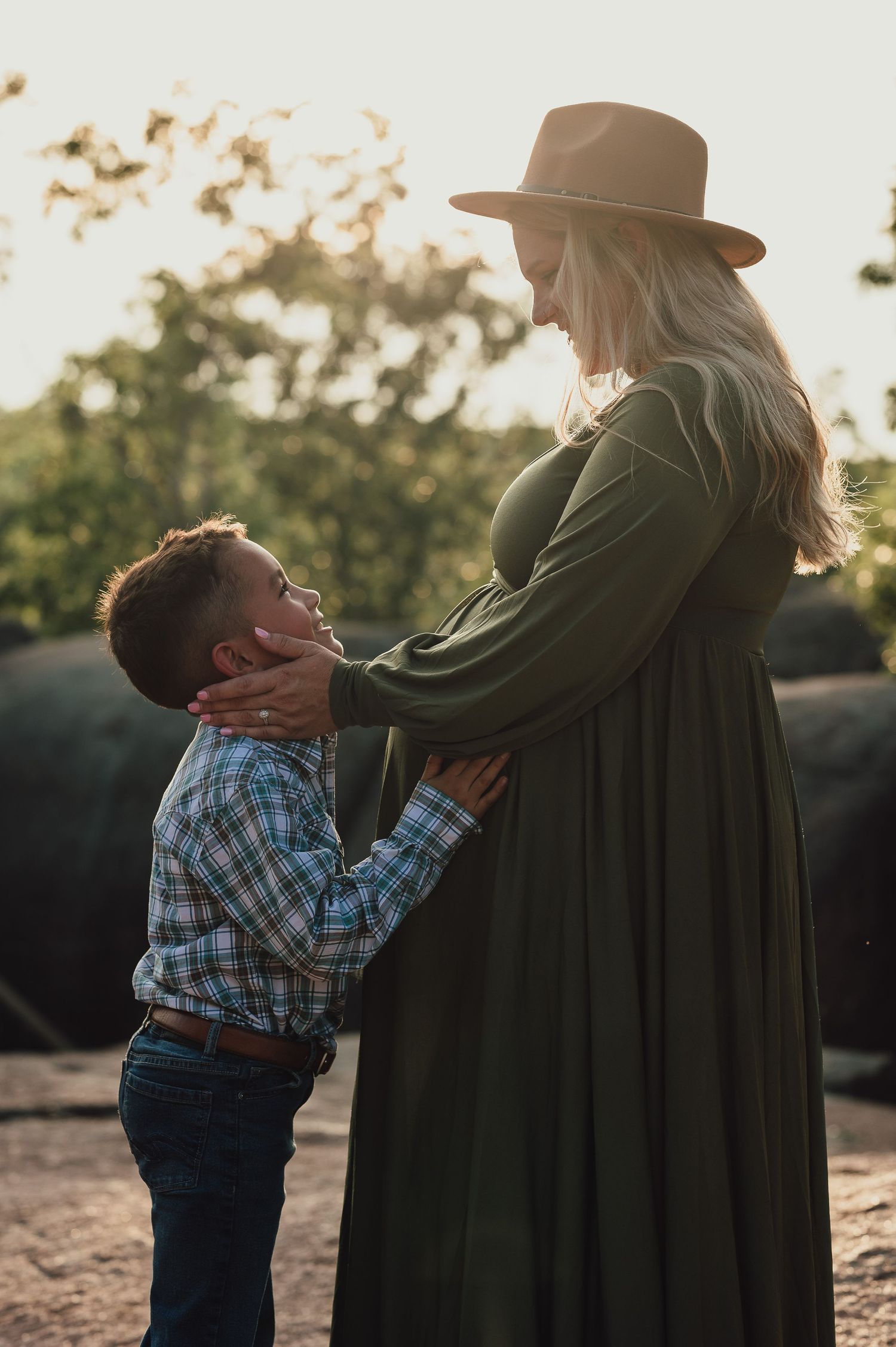 Sweet moment between child and expectant mother in olive dress during sunset photoshoot.