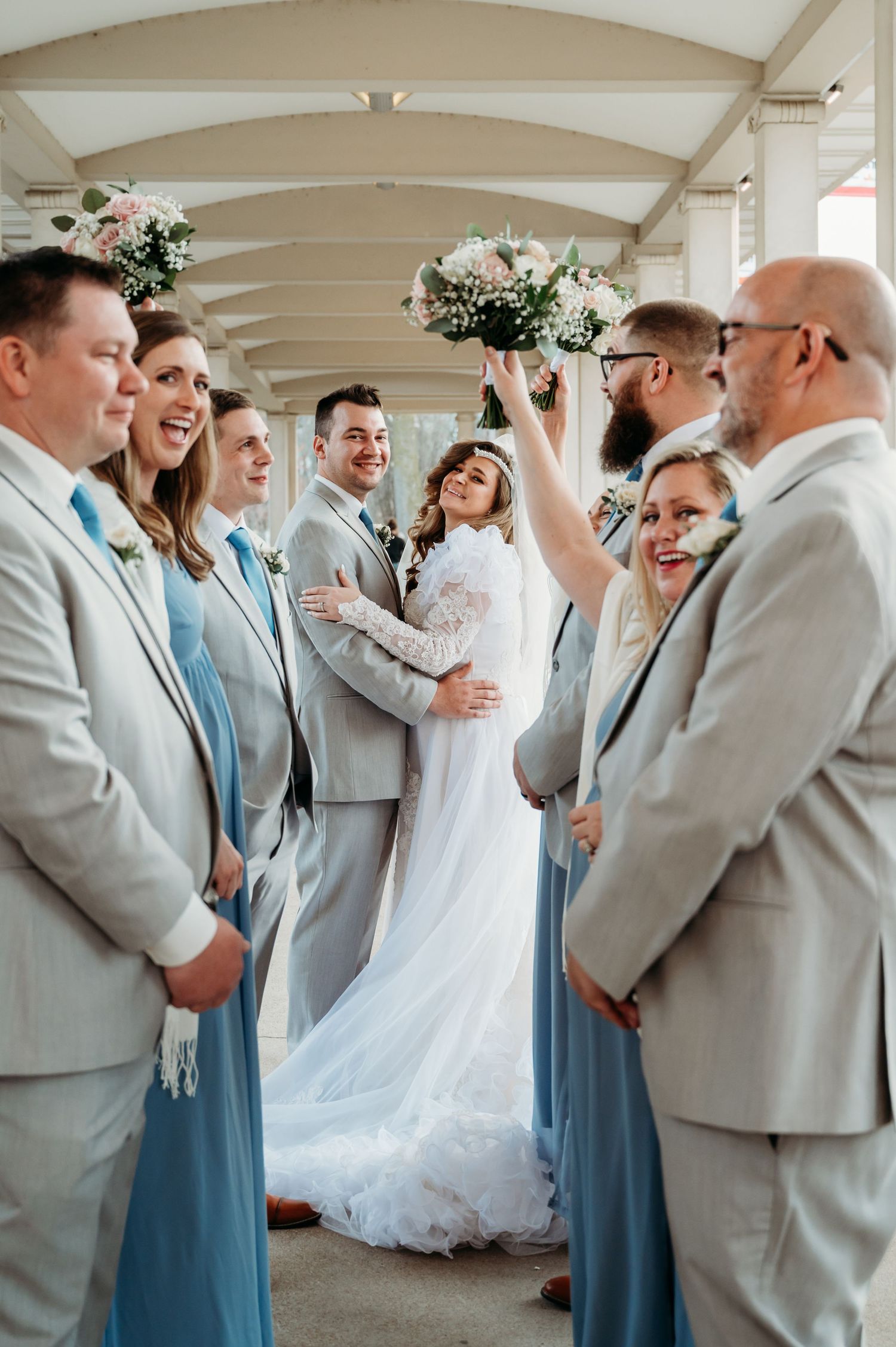 Wedding party members in light gray suits and blue dresses share joyful reactions during celebration moment.