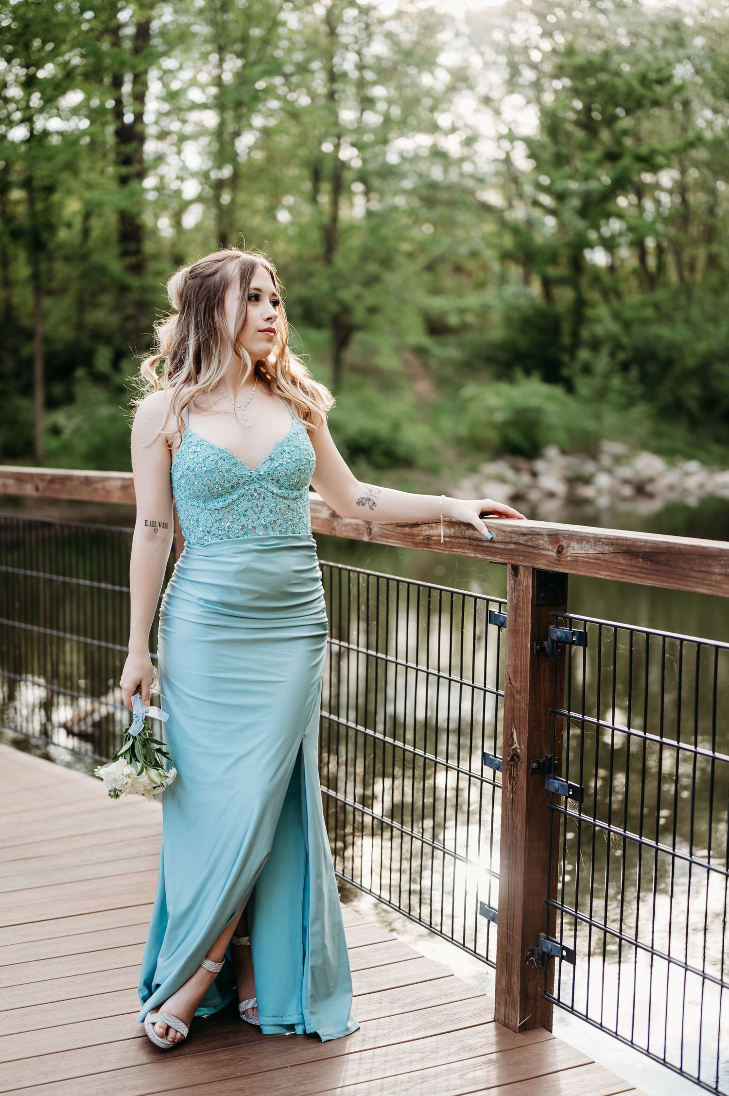 Prom girl in aqua dress poses elegantly on wooden deck overlooking water.
