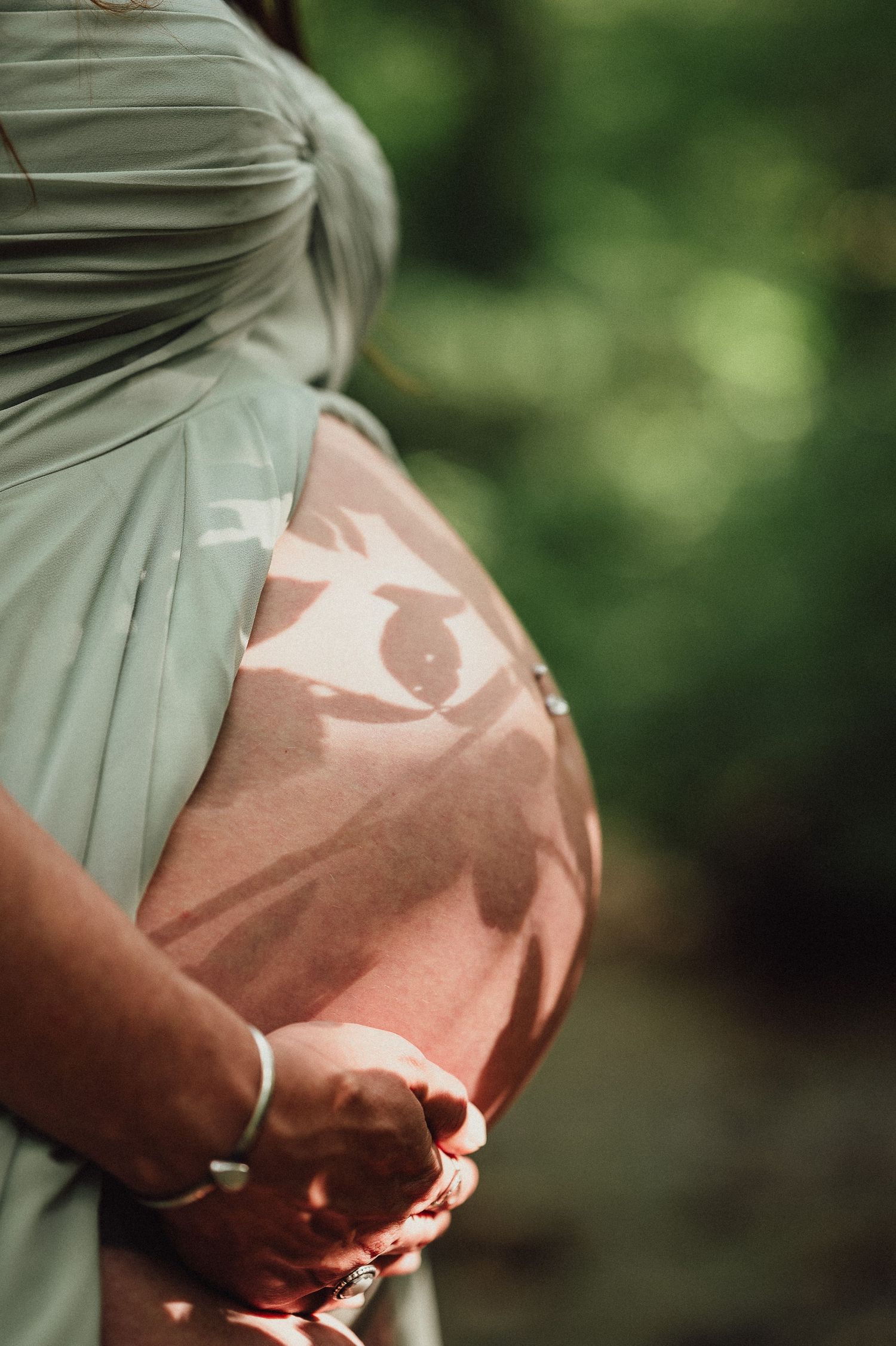 Close-up shots of pregnant belly in sage green dress with sunlight creating natural shadows.