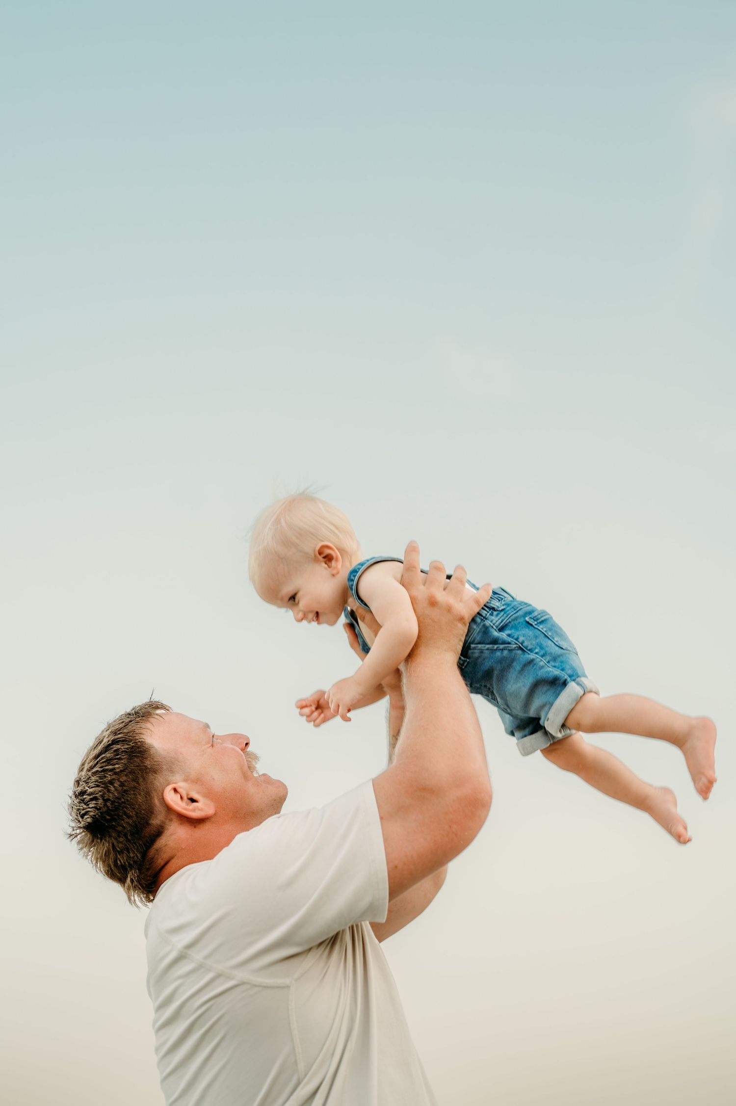 Playful moment captured of adult lifting small child against light blue sky background. Captured by Jessica Sagerty Studios