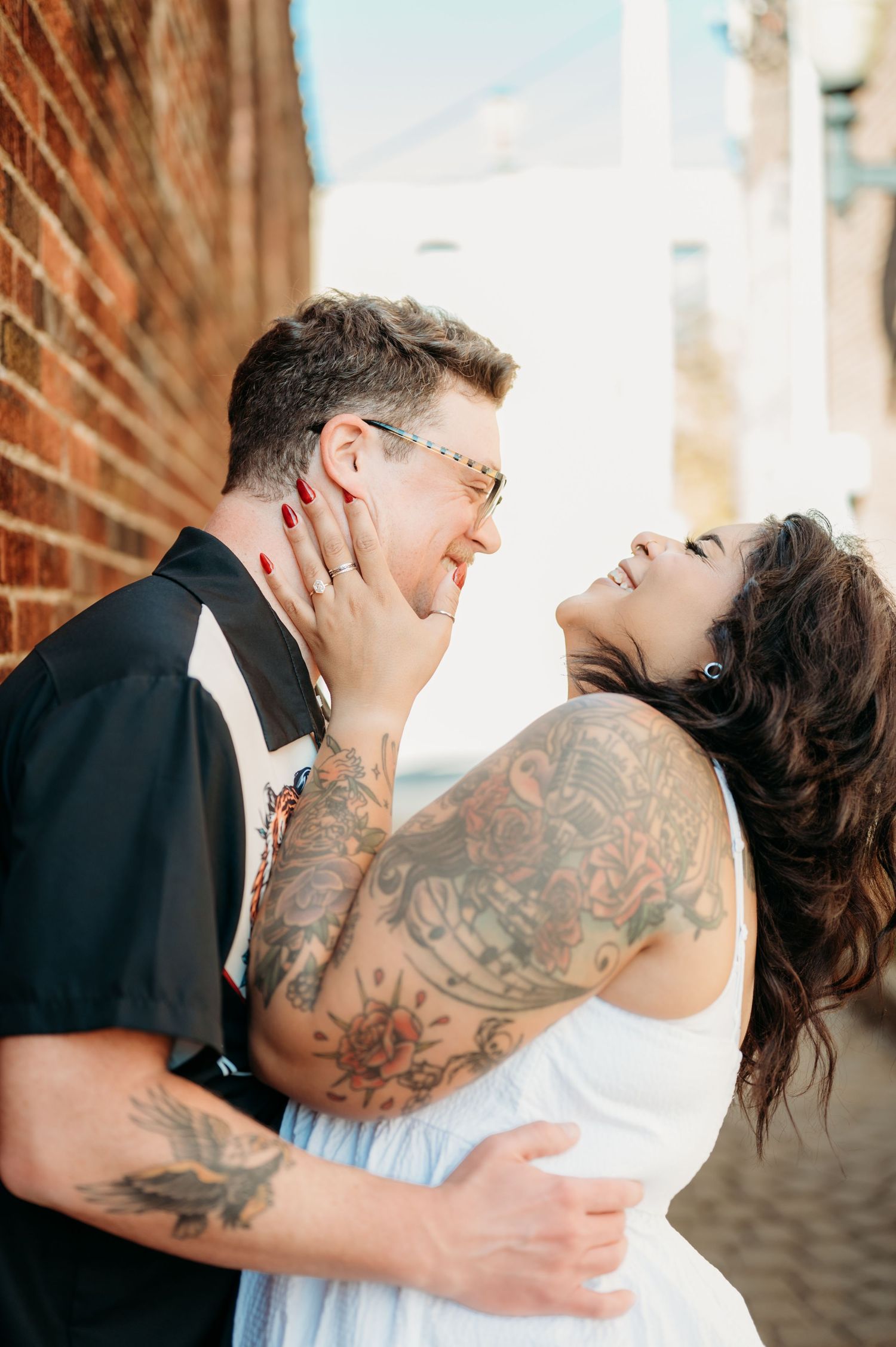 A joyful couple in wedding attire share a romantic moment against a brick wall, with visible tattoos on the bride's arm.