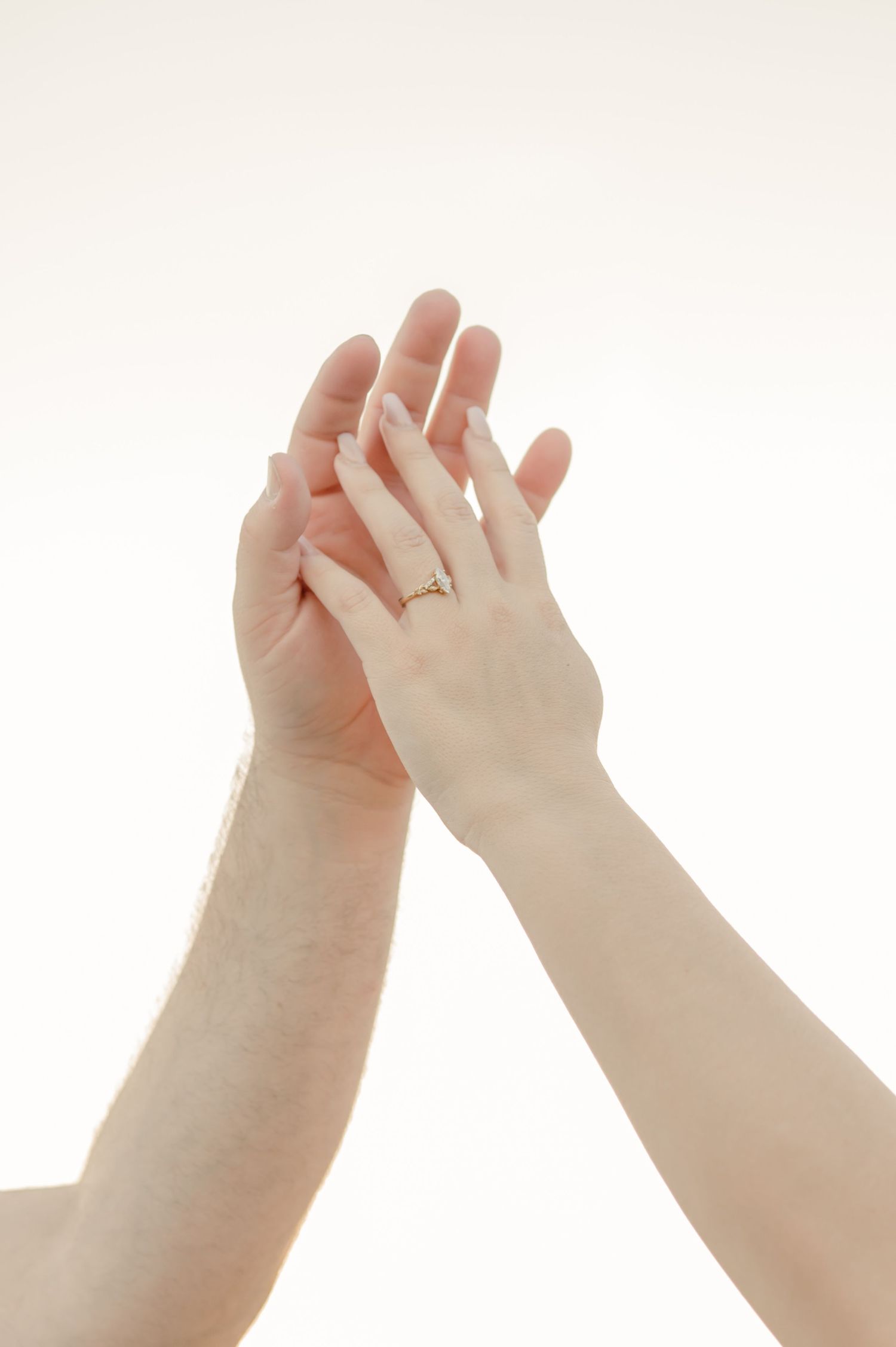 Two hands reaching upward against a light background with one hand wearing an engagement ring.