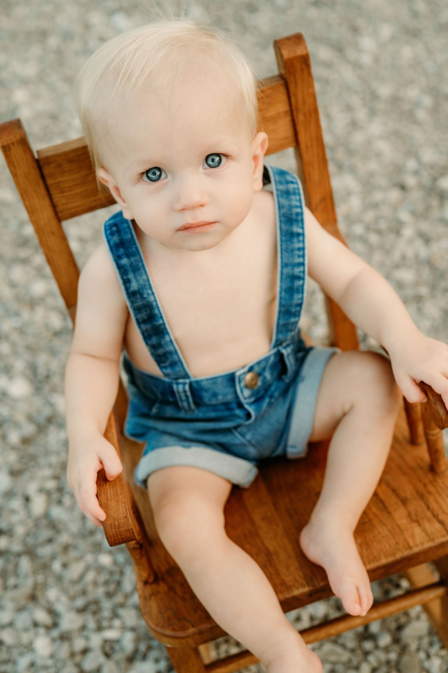 Small child wearing denim overalls sits in wooden chair on gravel surface.
