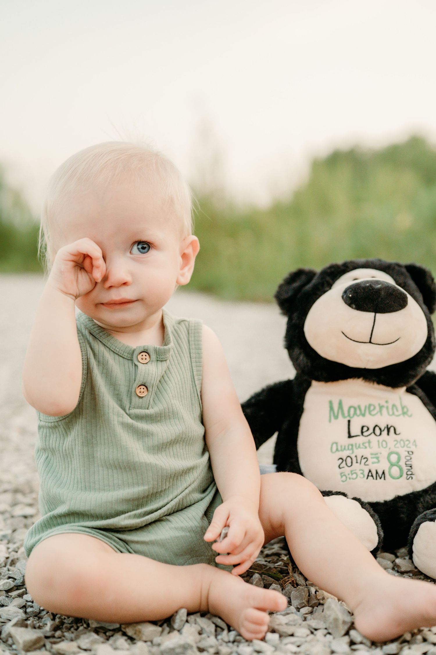 Baby in mint green outfit sits next to plush teddy bear on rocky ground. First birthday session