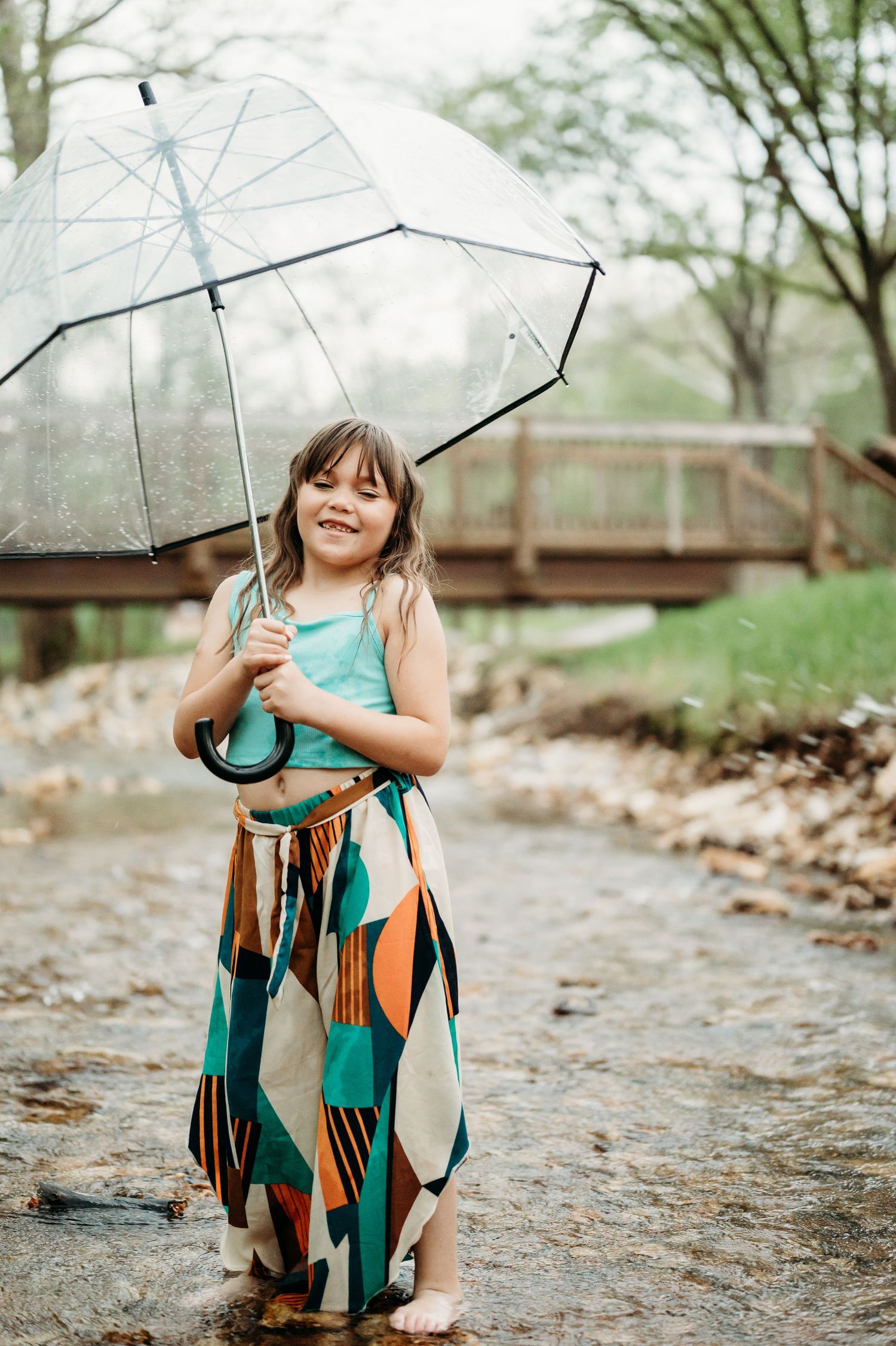 Child in turquoise shirt holding clear umbrella while playing in nature.