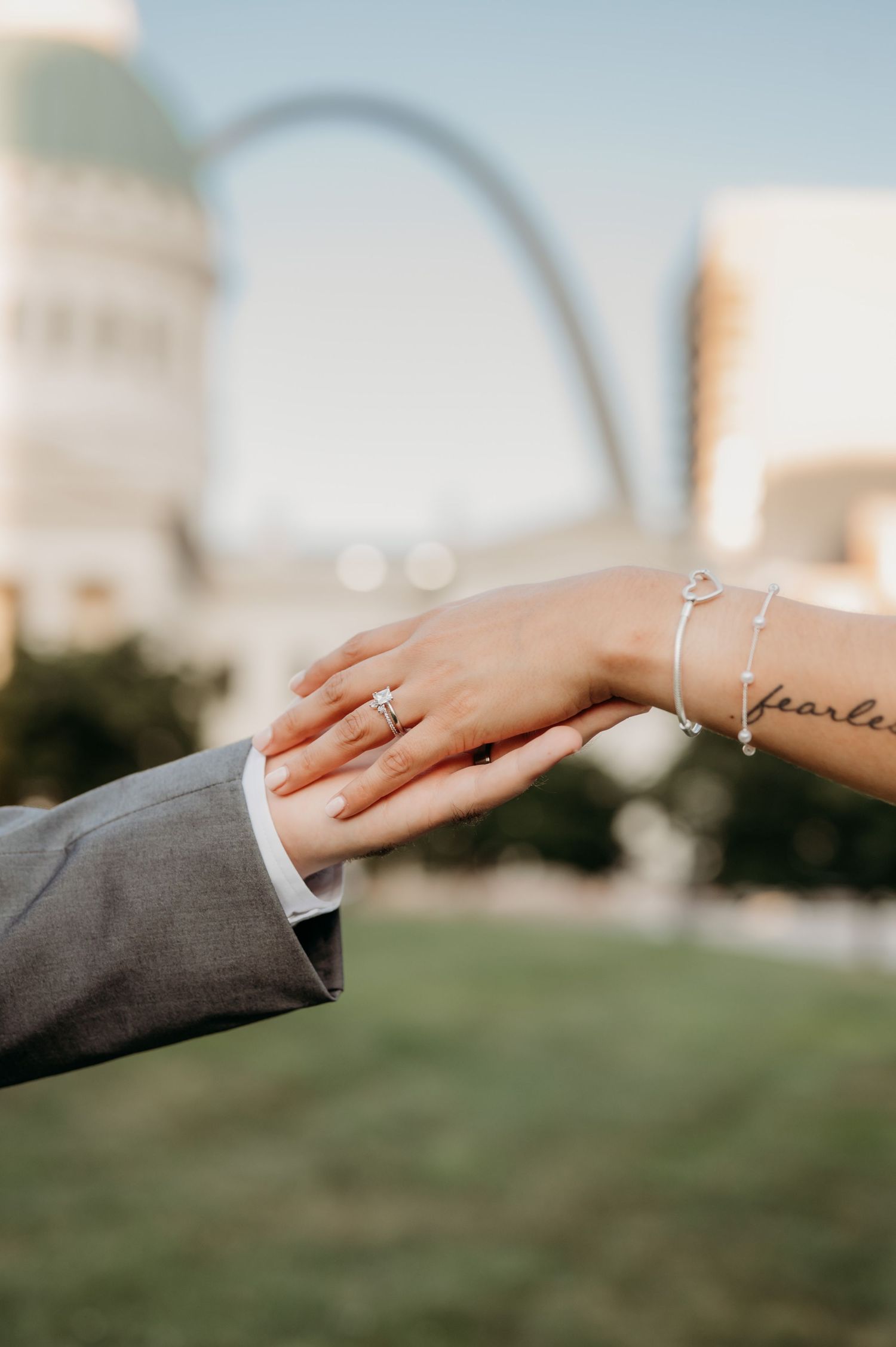 Hands with engagement rings touch tenderly against a blurred outdoor backdrop.