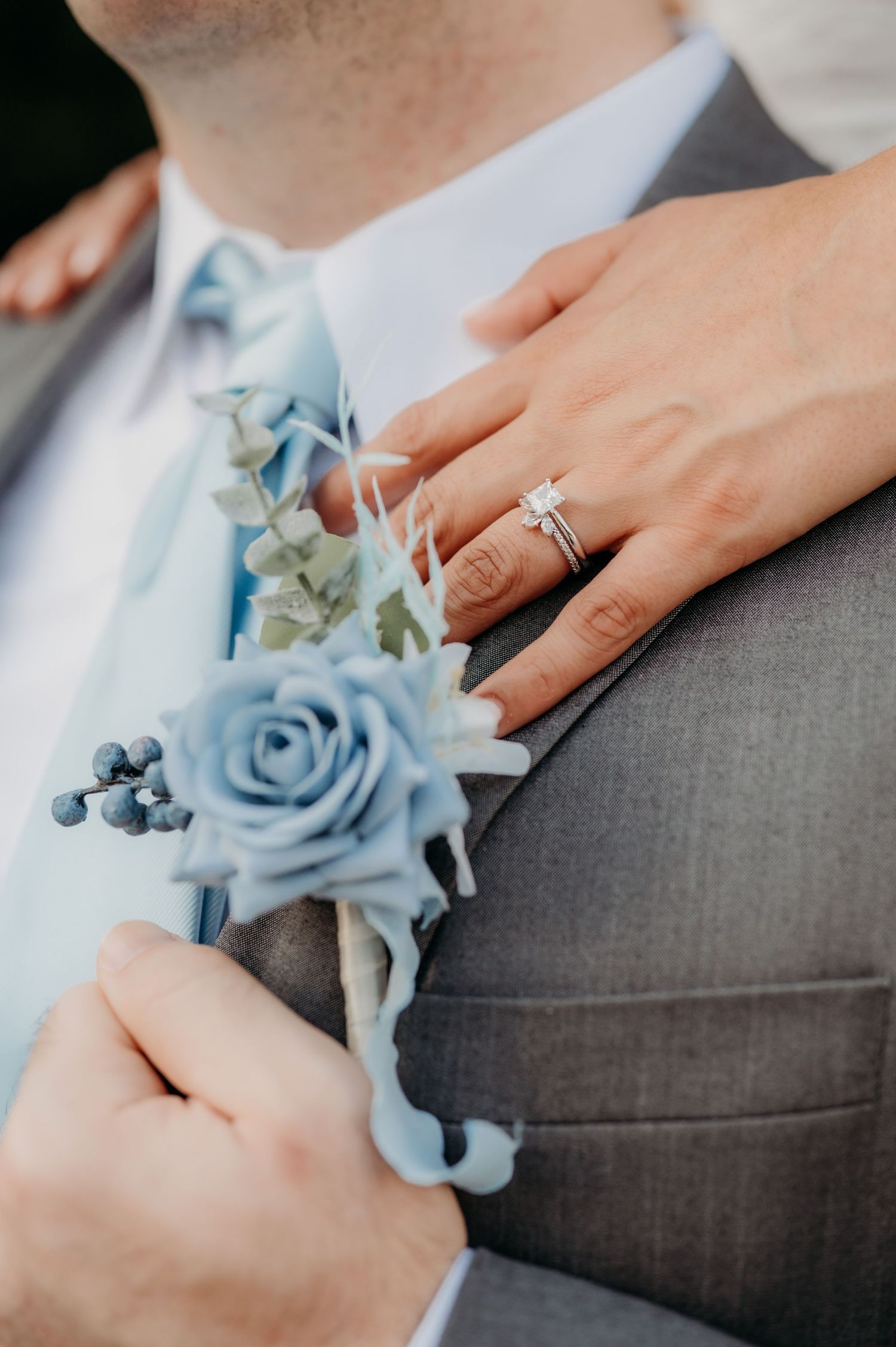 Hands pinning a light blue rose boutonniere onto a gray suit jacket at a formal event.