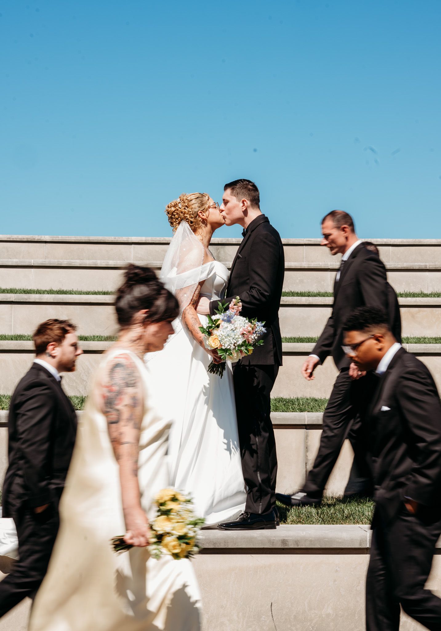 Bridal party walking in sequence against a concrete wall under bright blue sky, creating motion blur effect. Captured by Jessica Sagerty