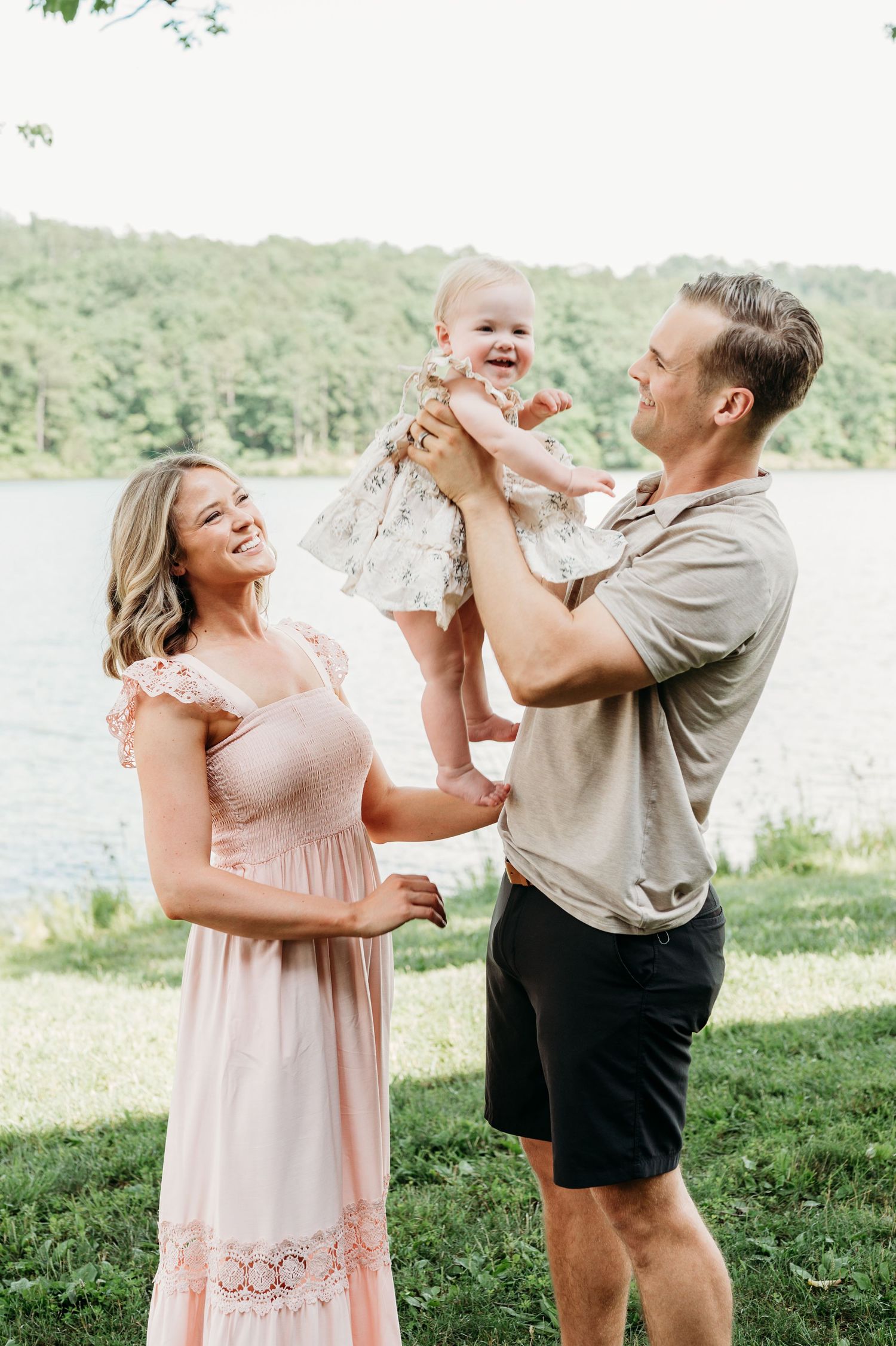 Family enjoying playful moments together on a grassy hillside near a lake in matching pink and neutral outfits.