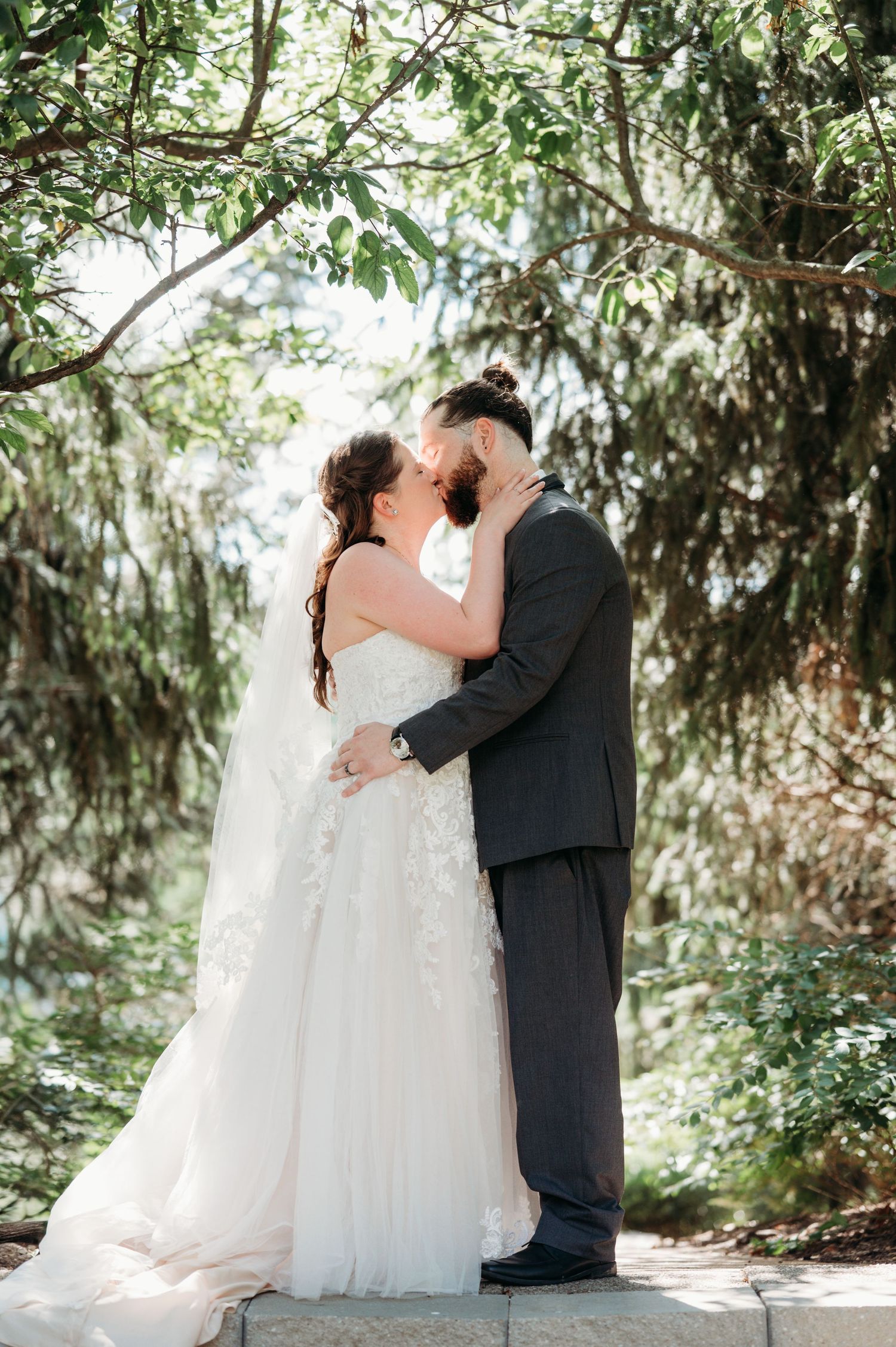 Romantic wedding couple embracing under trees in a sunlit garden setting.