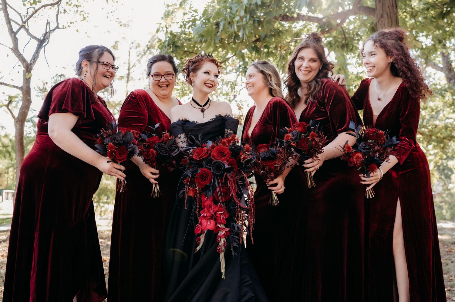 A group in burgundy velvet dresses and black lace stand together holding red and black floral bouquets outdoors.